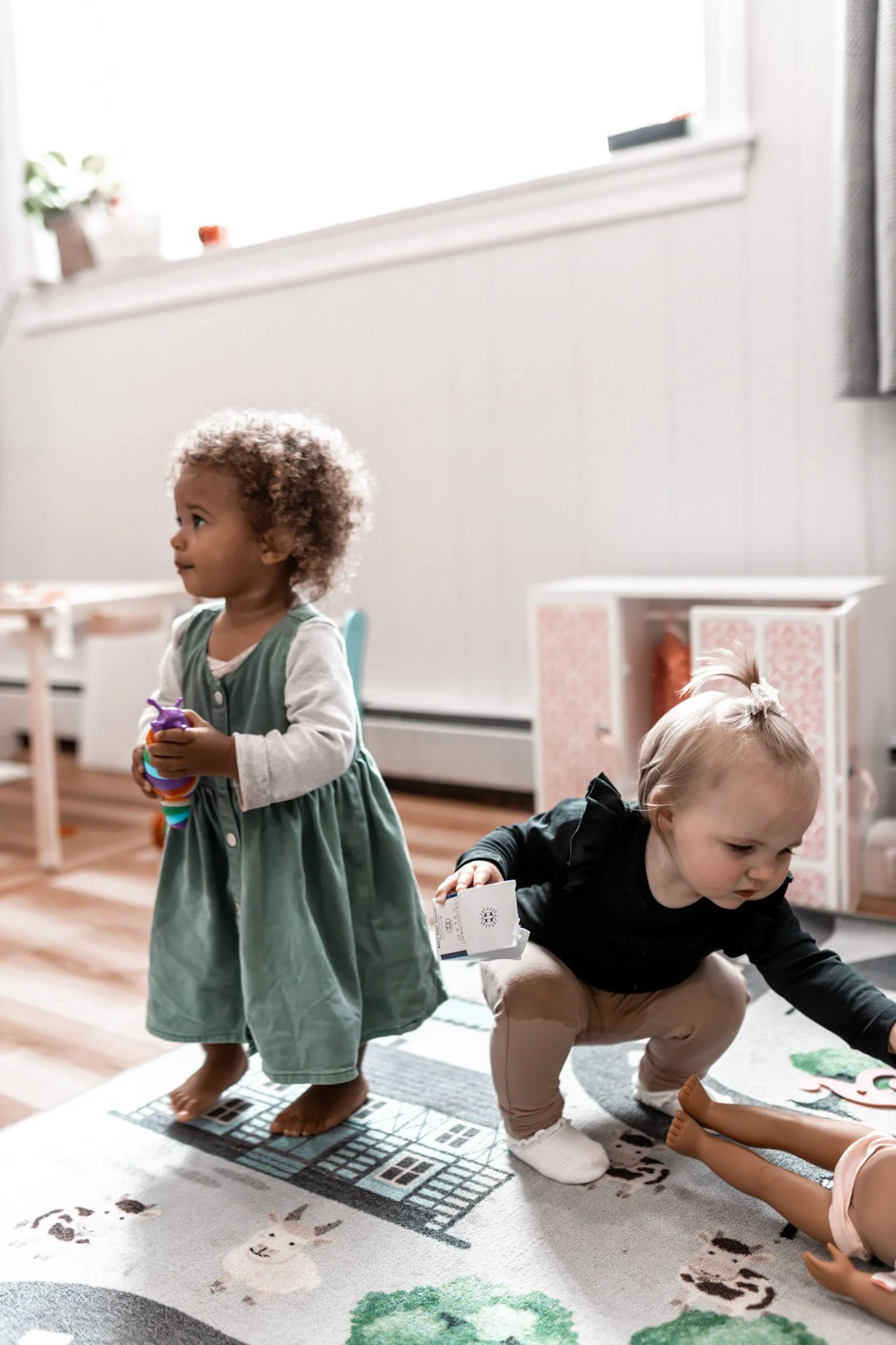 Two young children playing on a colorful rug in a bright room. One girl with curly hair wearing a green dress stands holding toys, while another girl with blond hair in a ponytail and a black shirt crouches reaching for a doll's feet.