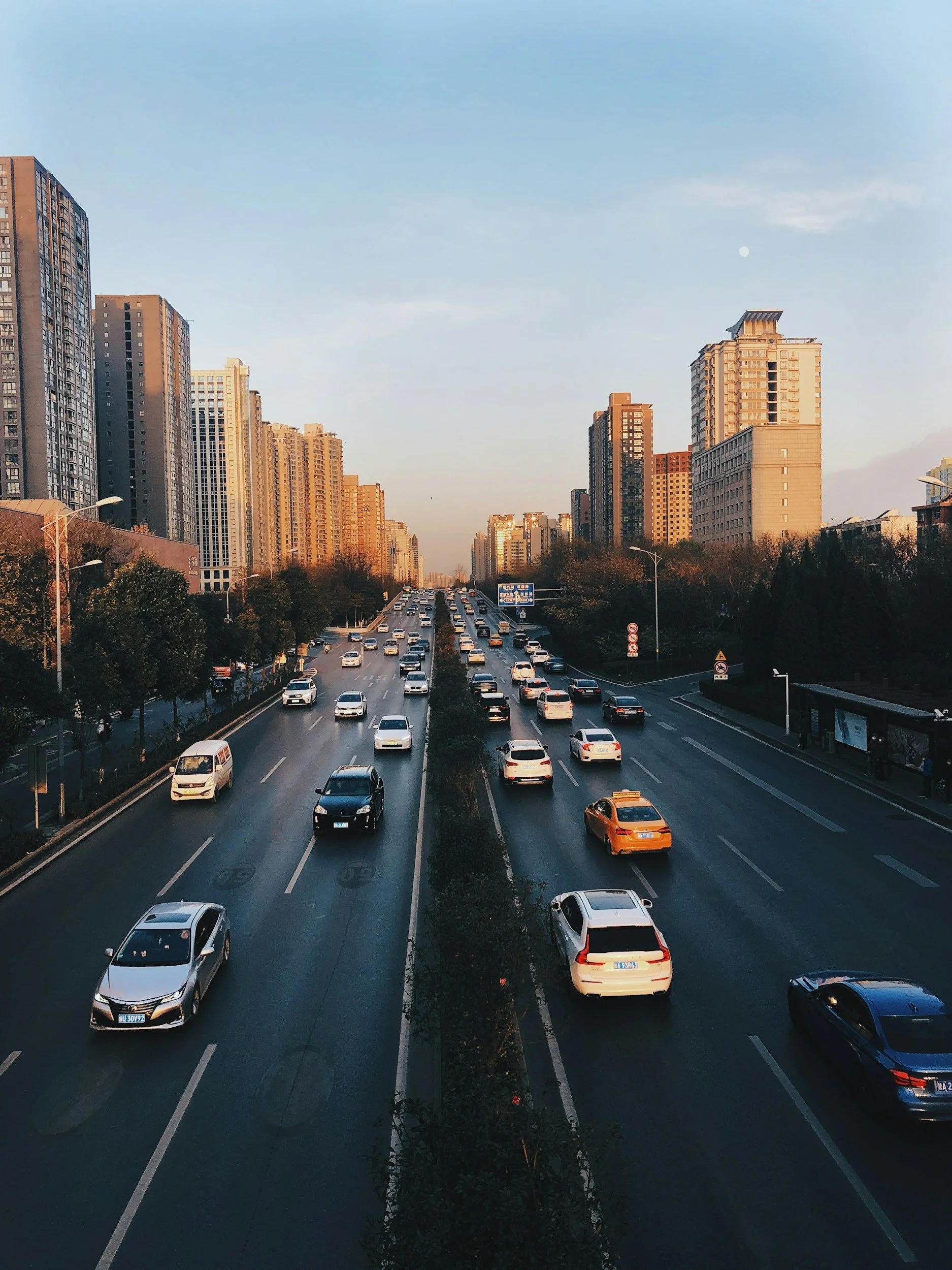 City street with multiple lanes of moving cars and tall buildings on both sides during sunset.