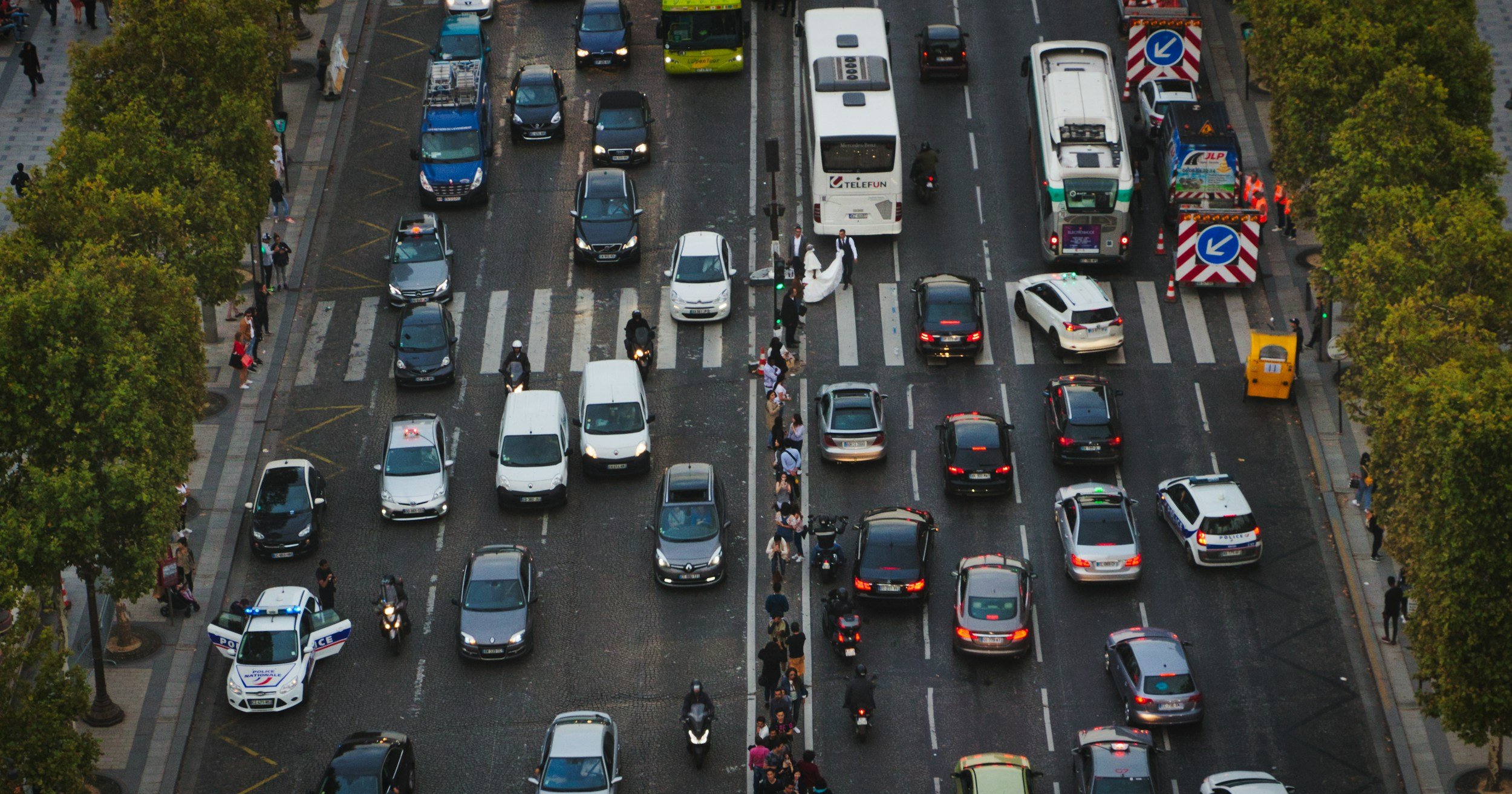 An aerial view of a busy city street with multiple lanes of traffic, including cars, buses, motorcycles, and police vehicles. Pedestrians are walking on sidewalks on both sides of the street, and trees line the edges of the sidewalks.
