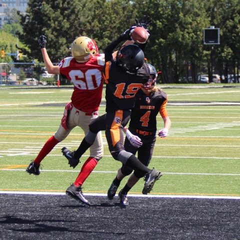 Football players in action during a game on a football field, with one player in a red and beige uniform jumping and another in a black and orange uniform catching the ball.