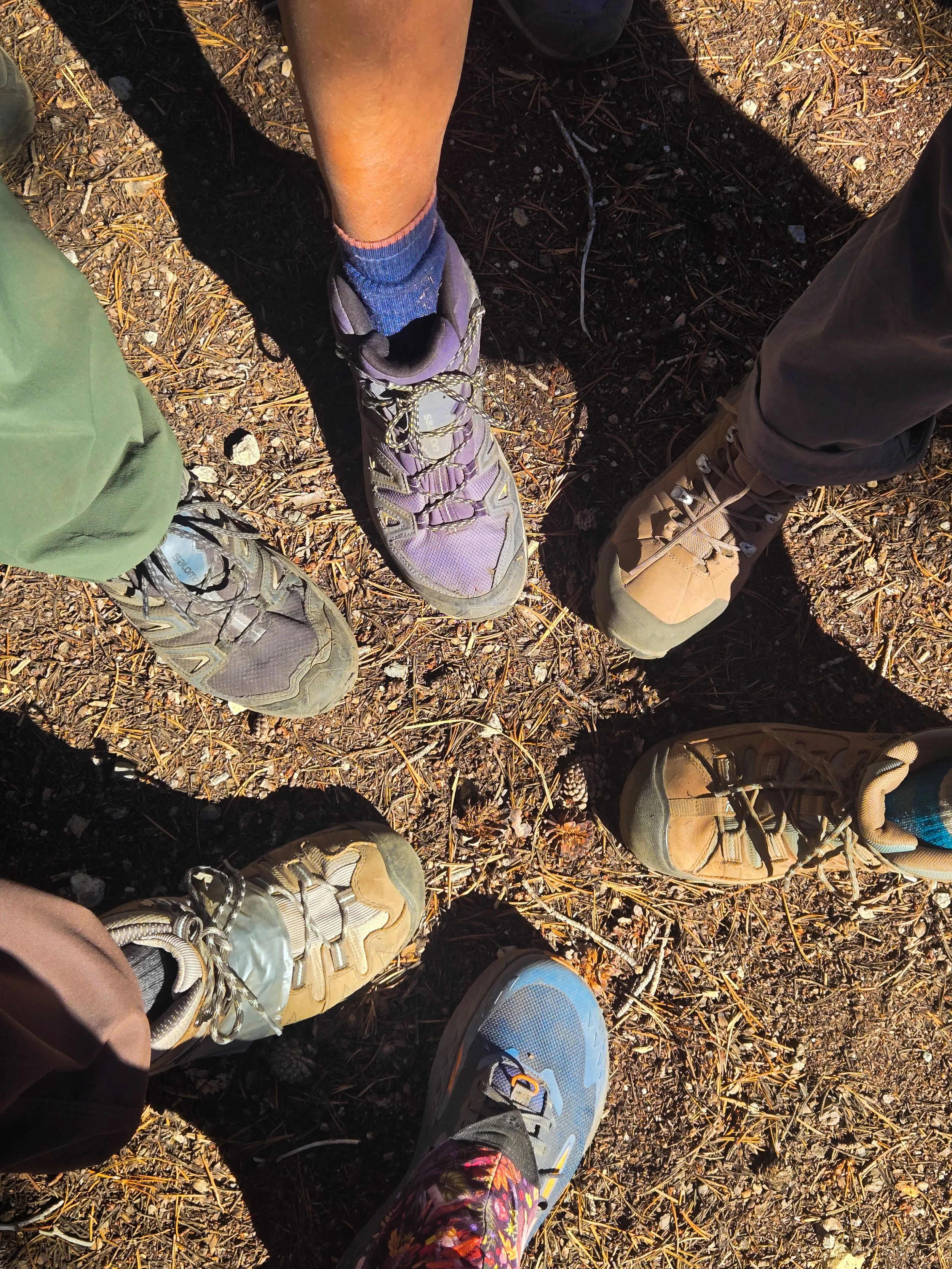 Six backpackers standing in a circle outdoors, showing their hiking shoes on a dirt trail. The circular formation of hiking boots suggests unity, comradery, together as a team.
