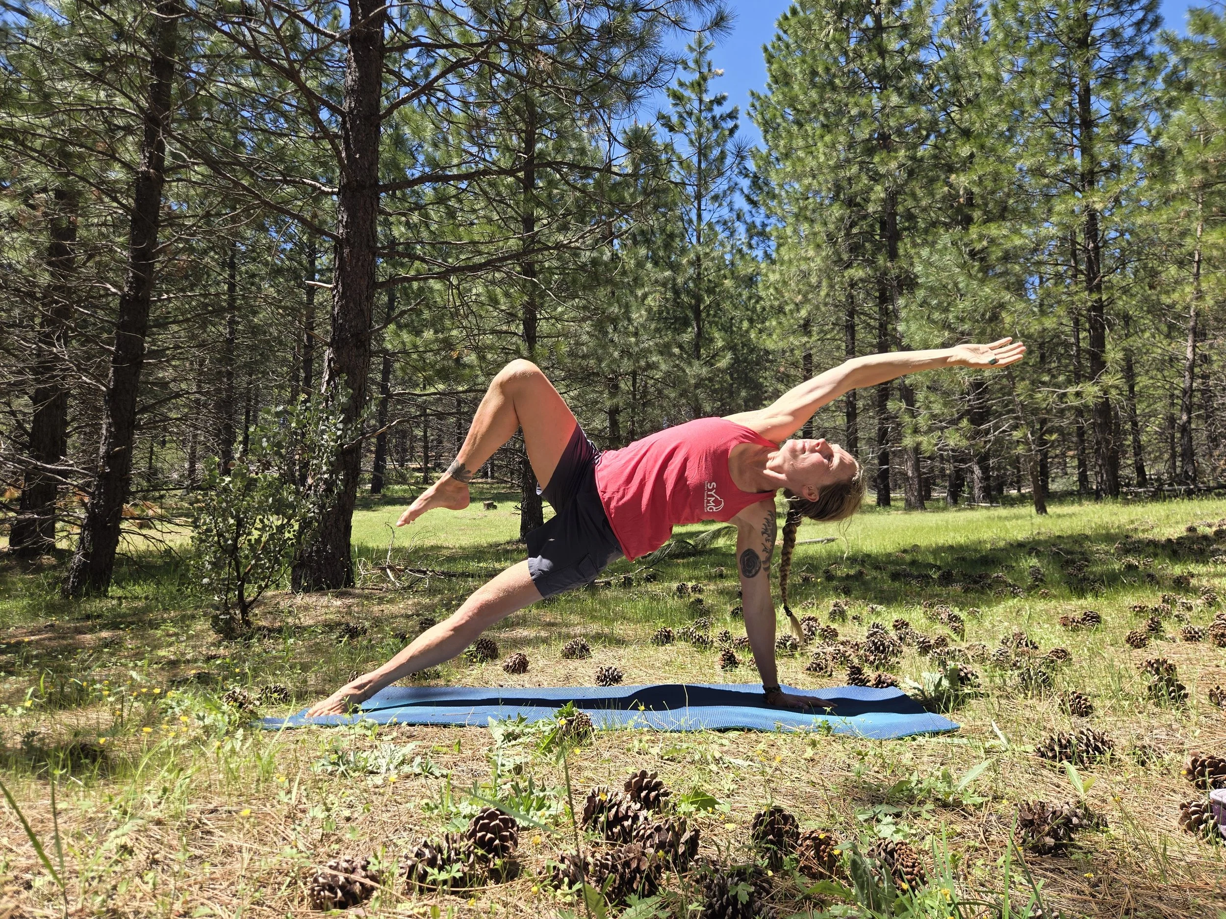 A woman practicing yoga outdoors in a forest, performing a side plank pose on a blue mat amidst pine trees and pinecones.