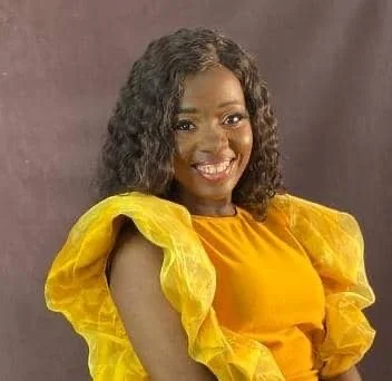 A young woman with shoulder-length black hair smiling outdoors among green plants.