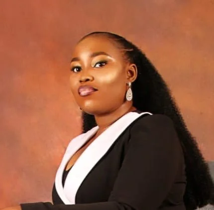 A close-up of a man with curly brown hair, glasses, and a beard, sitting indoors with a blurred background that includes a plant.