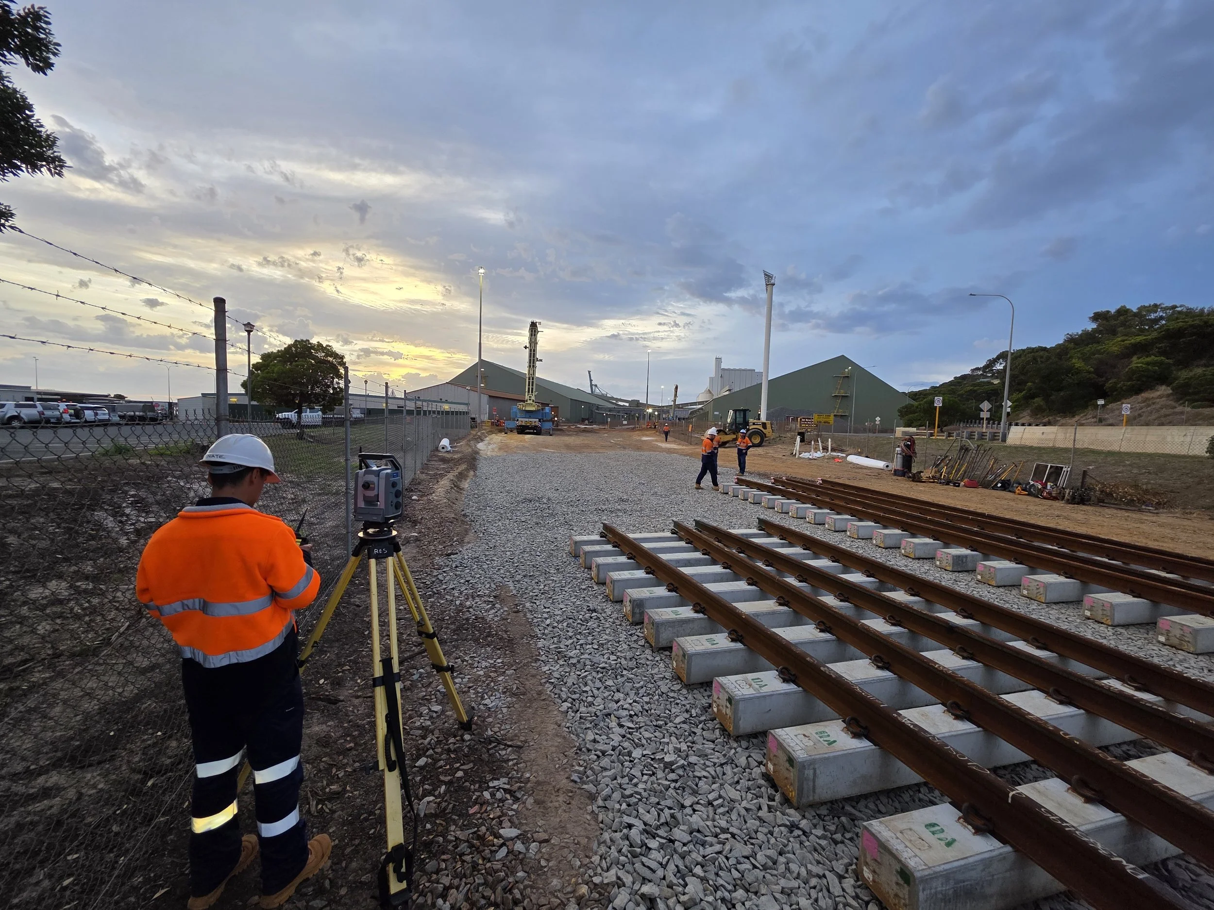 Construction workers at a railway site during sunset, with tracks being installed and heavy machinery in the background.
