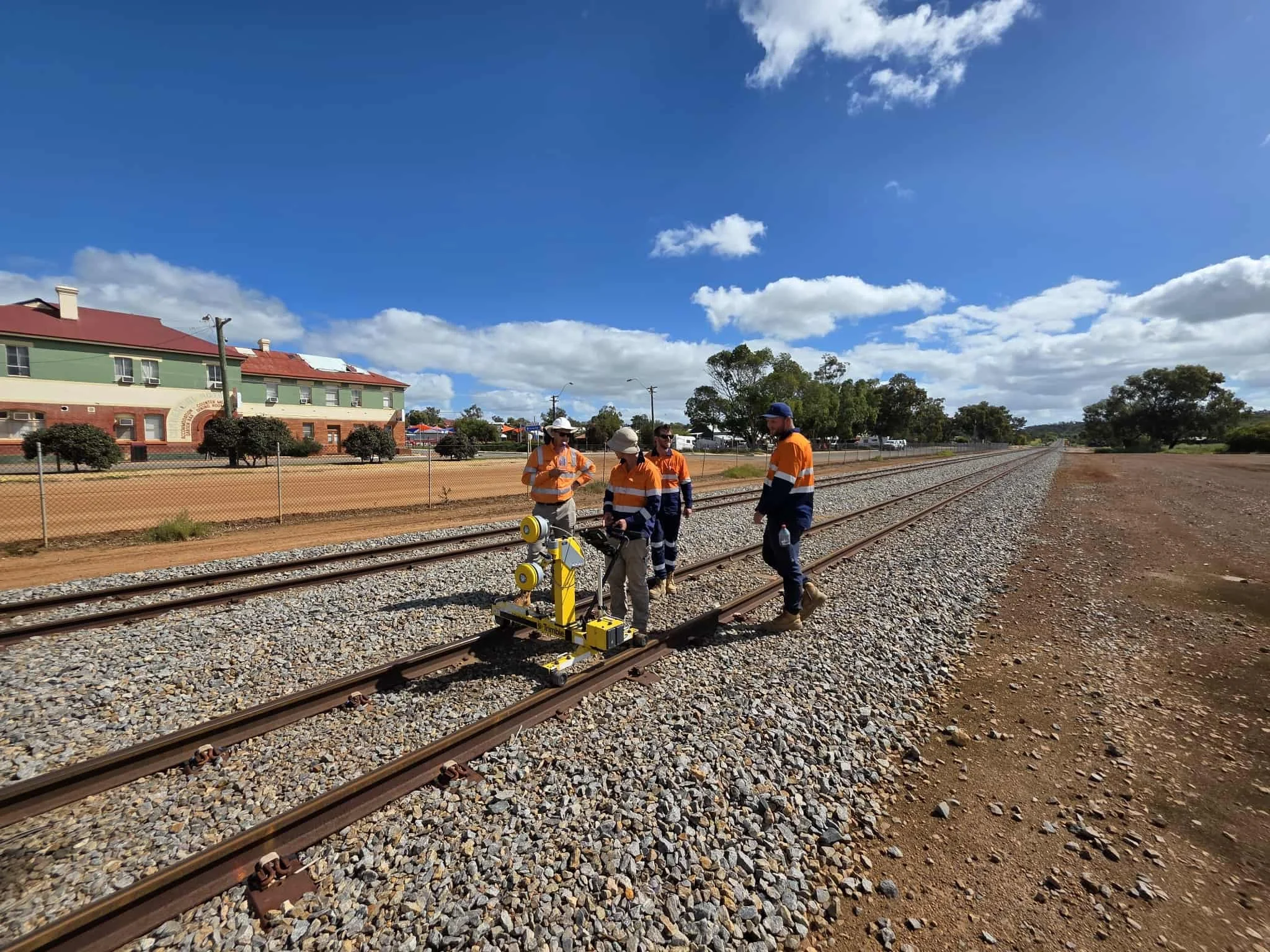 Group of four railway workers, wearing high-vis safety clothing and hats, inspecting and working on train tracks under a partly cloudy sky.