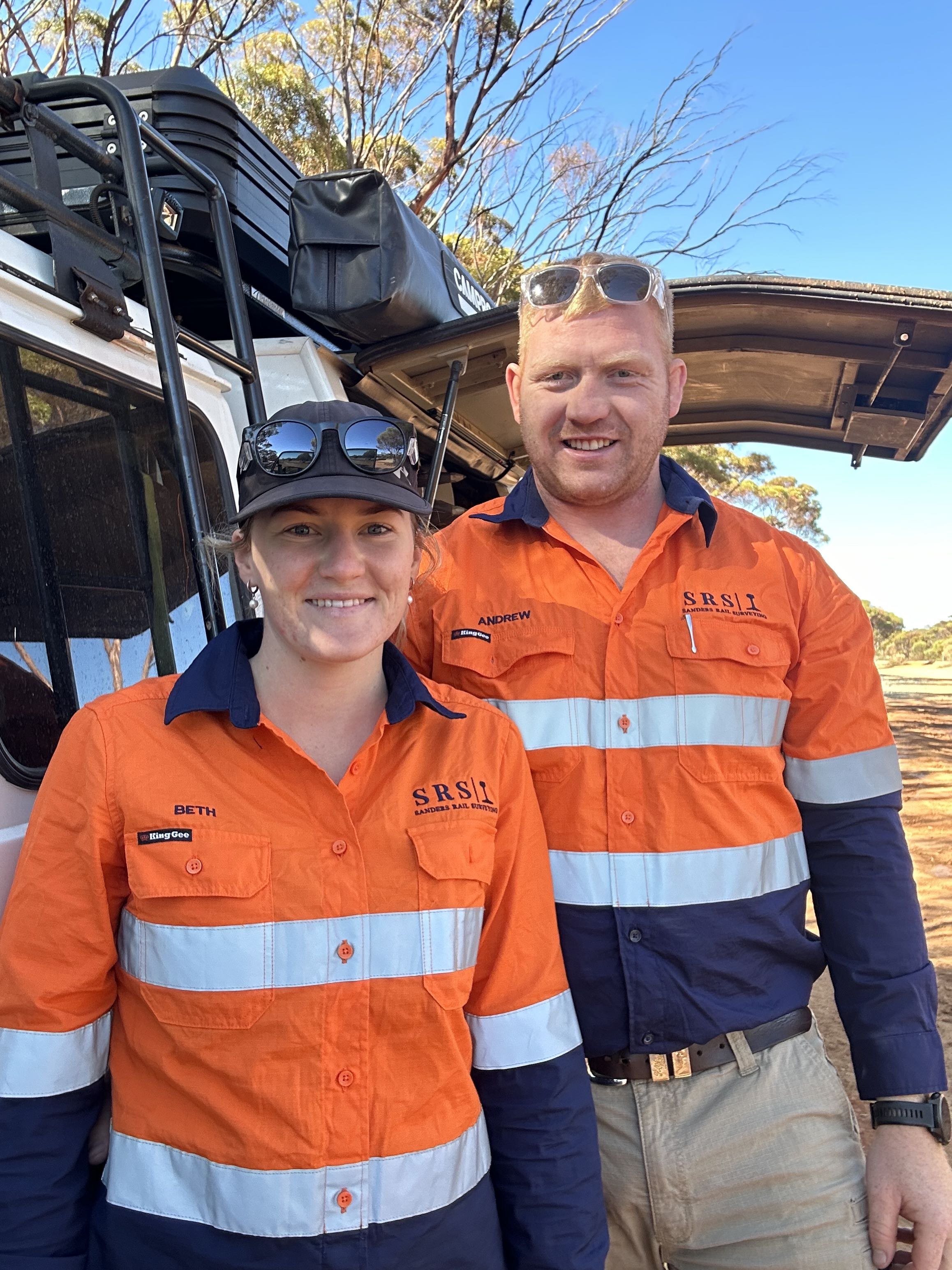 Two outdoor workers in orange safety shirts with reflective stripes, standing in front of a vehicle with equipment on top, smiling at the camera.