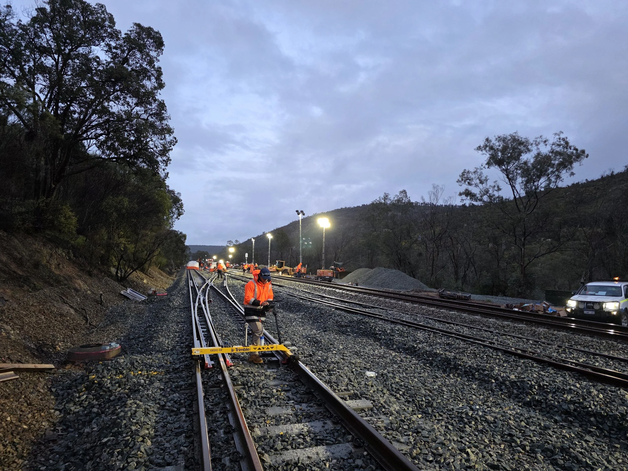 Workers in orange safety vests installing or repairing railway tracks during dusk, with construction equipment and lights in the background.