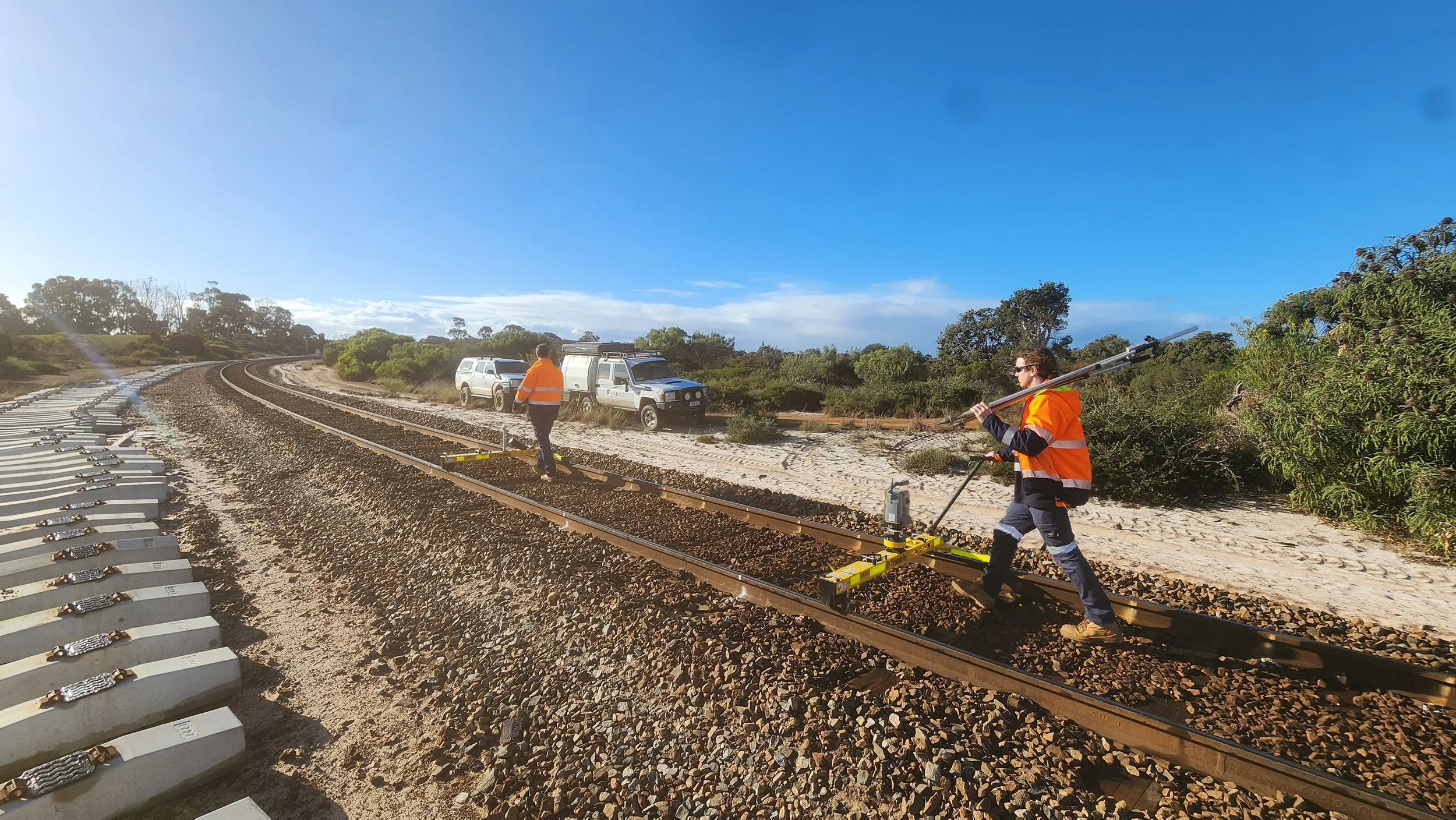 Two workers in orange safety vests installing or repairing railroad tracks along a sunny, rural railway with vehicles parked nearby and green trees in the background.
