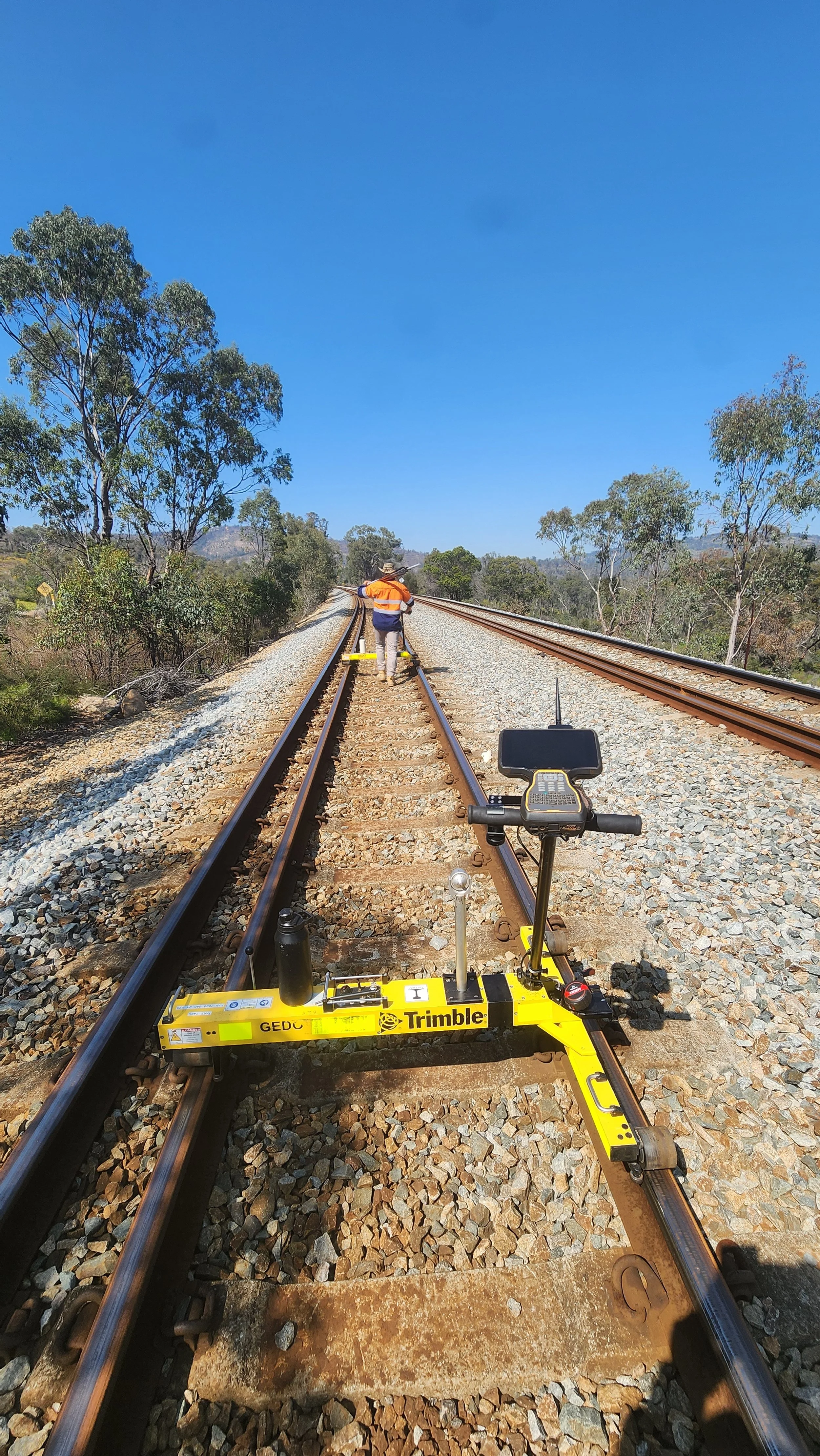 Track inspector in orange safety vest and hat on train tracks with surveying equipment and a robotic device in foreground, surrounded by trees and a clear blue sky.
