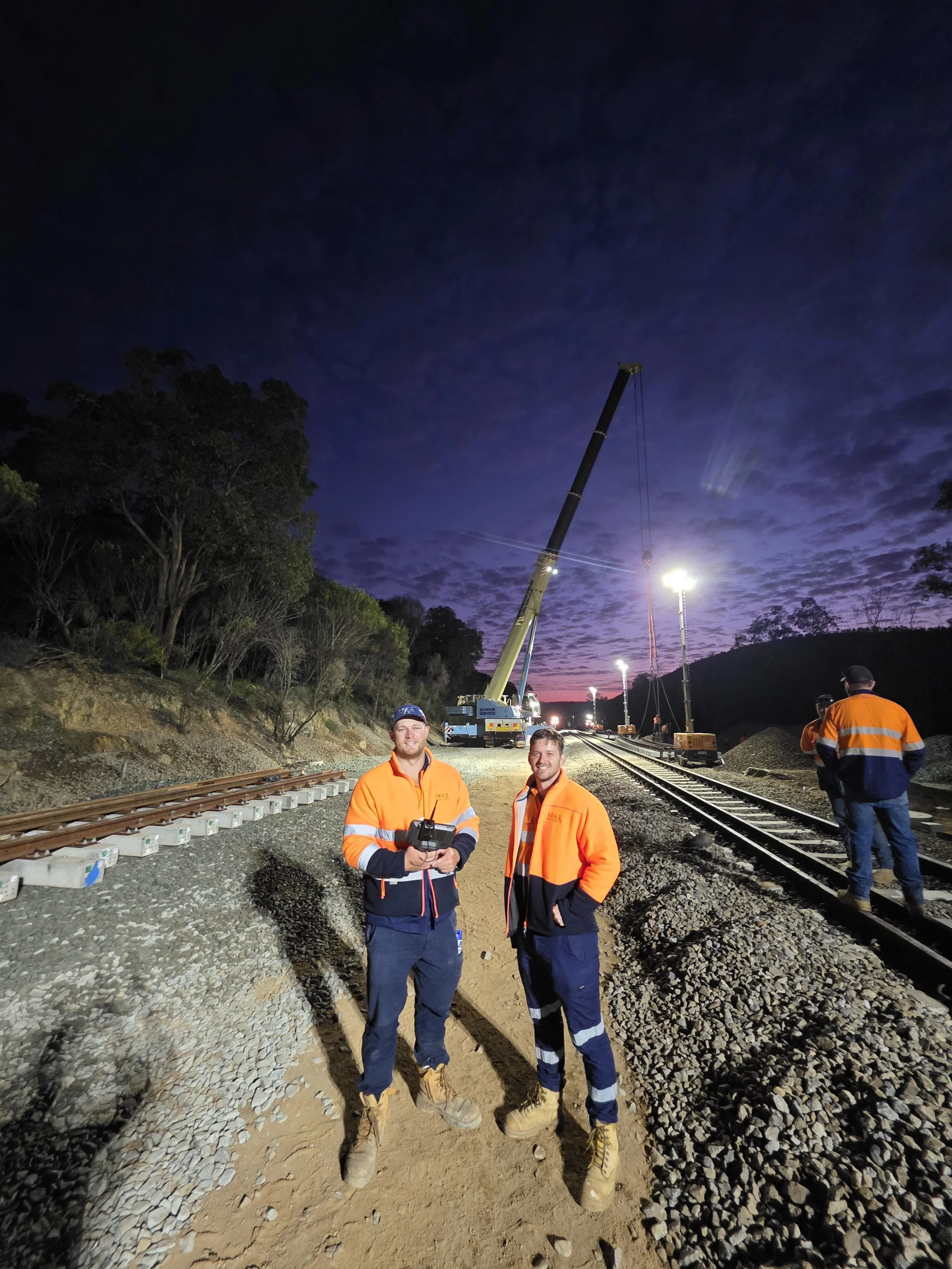 Two construction workers in orange and navy uniforms standing on a railway track at dusk, with a crane lifting equipment and a purple sky in the background.