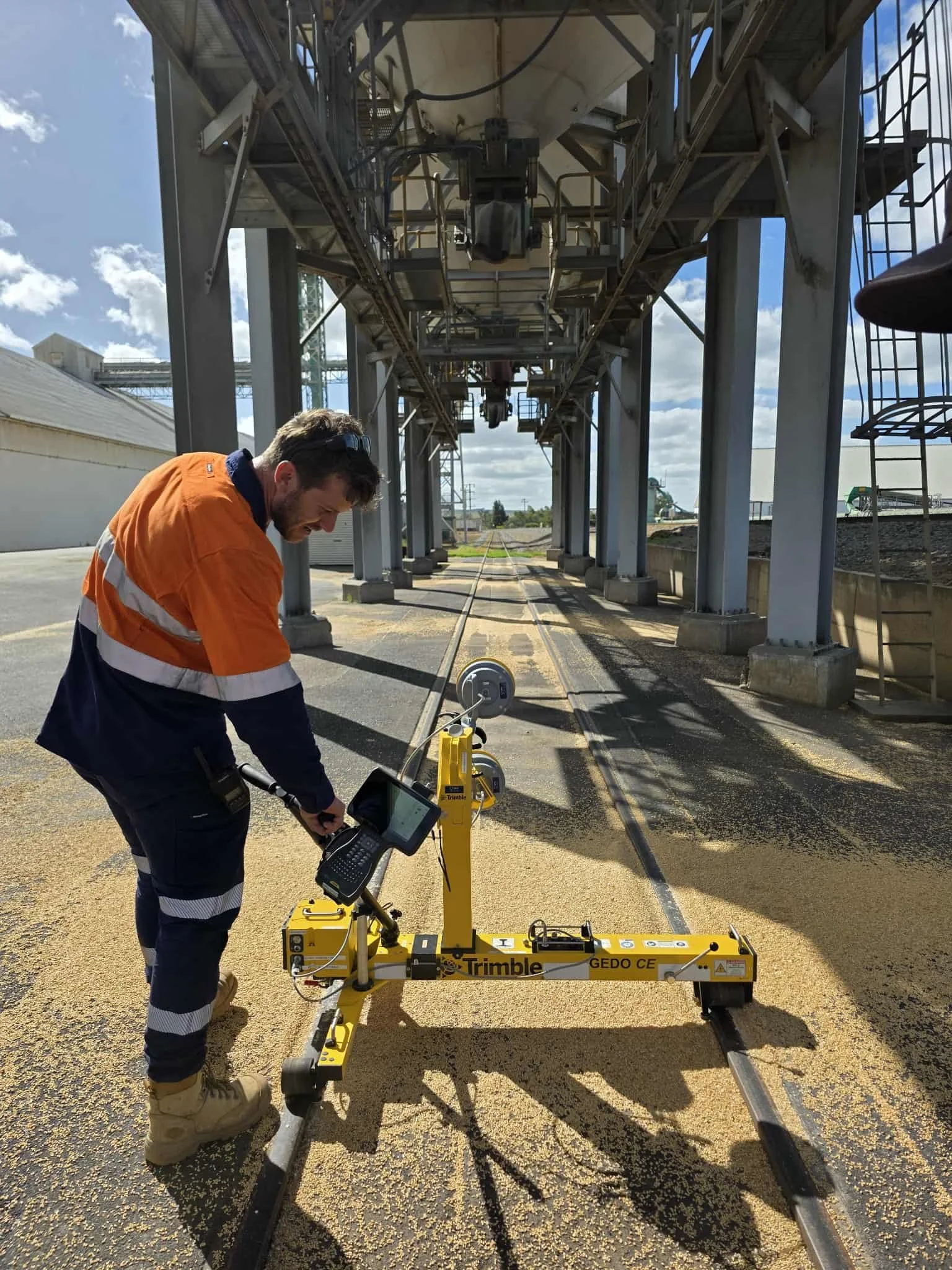 A worker wearing an orange safety vest and tan work boots uses a robotic device on railroad tracks outdoors under a metal structure, with a partly cloudy sky in the background.