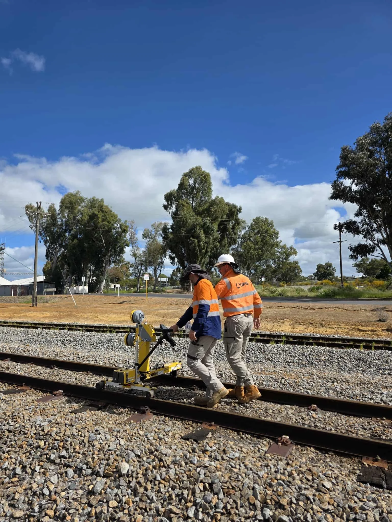 Two workers in orange and blue safety uniforms inspecting or working on a railway track with equipment, in a rural area with trees and power lines under a partly cloudy sky.
