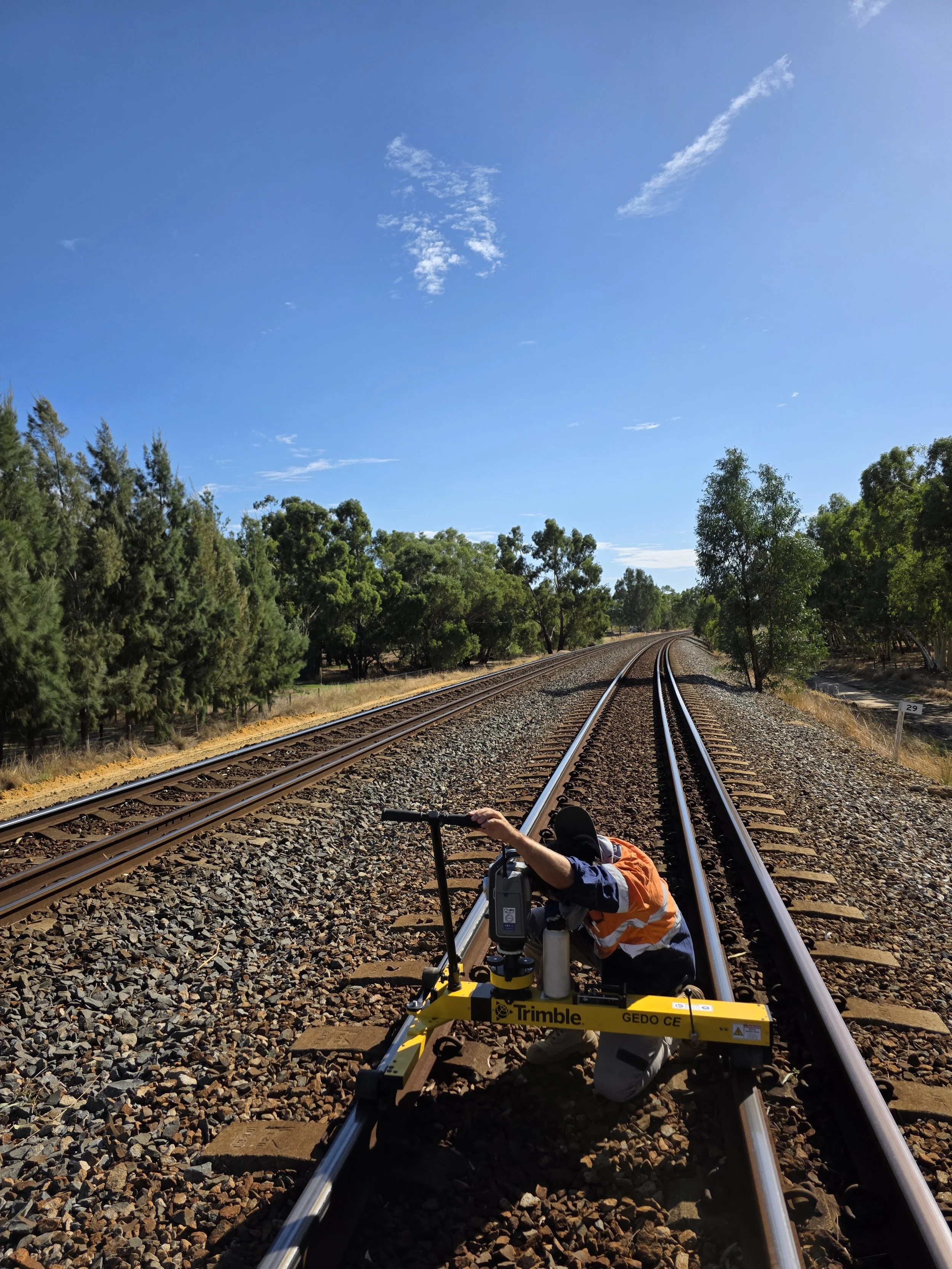 A person wearing a safety vest kneeling on railroad tracks while operating surveying equipment under a clear blue sky.