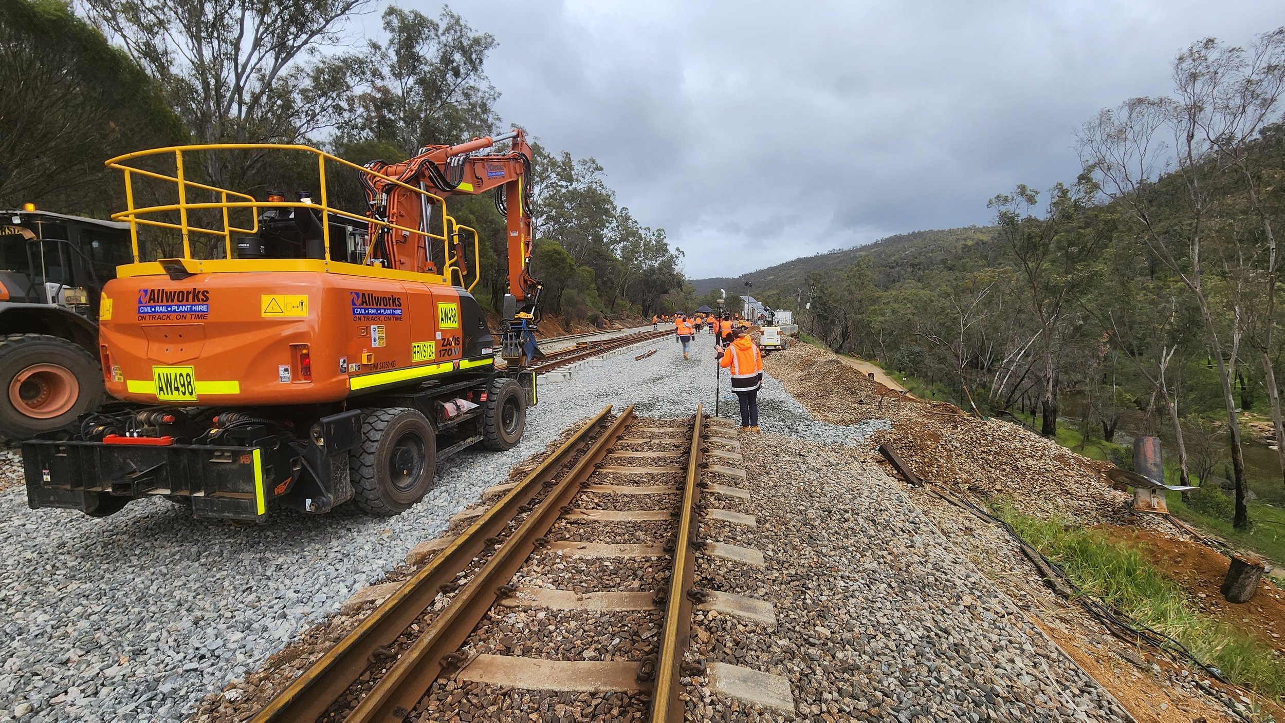 Workers in orange safety vests and helmets repairing and maintaining railroad tracks in a rural area with trees and hills in the background, and cloudy sky overhead.