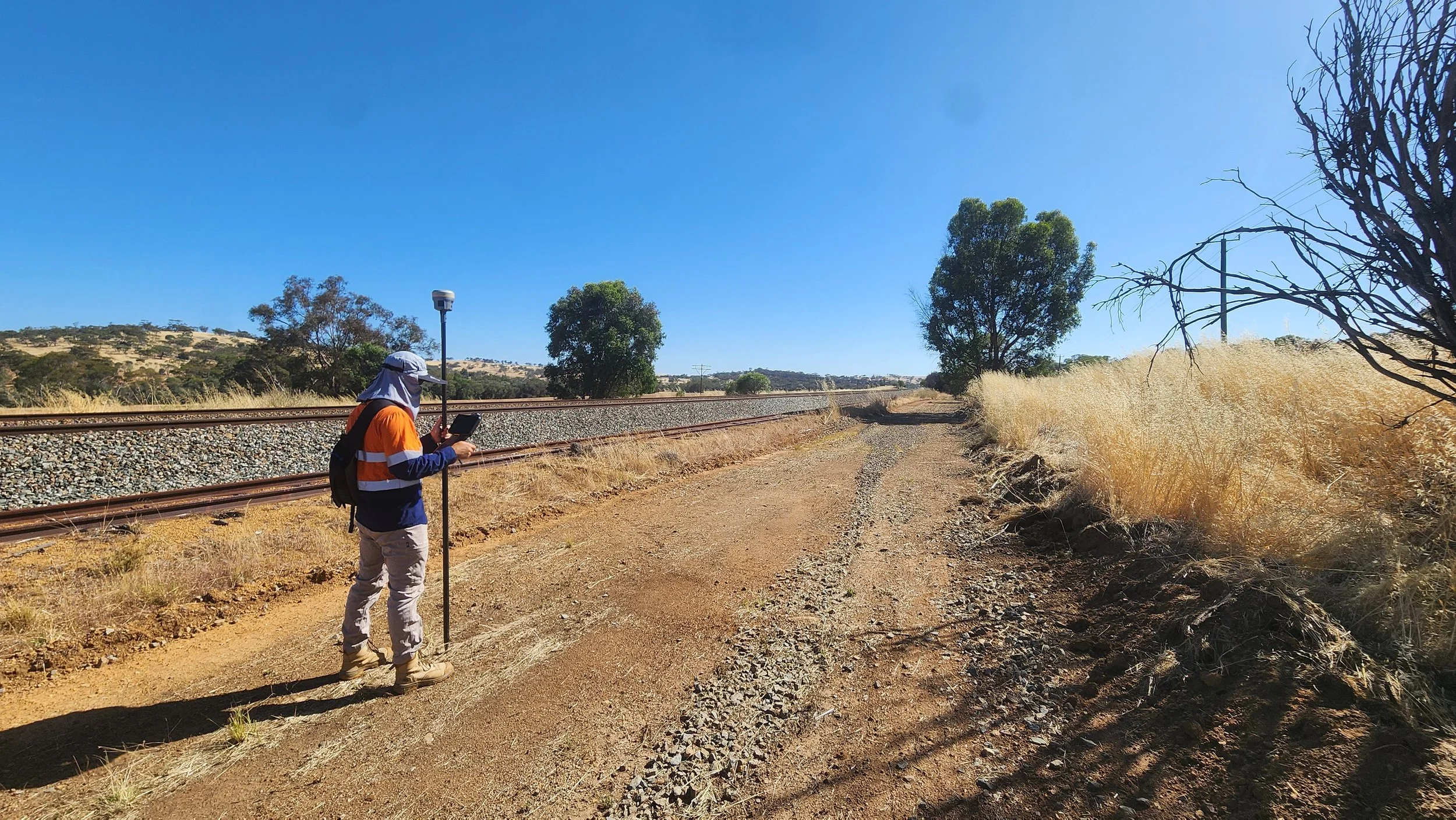 A person wearing an orange, blue, and white high-visibility jacket, beige pants, tan boots, and a hat, standing on a dirt path beside a railroad track, looking at a tablet. The scene is sunny with a clear blue sky, dry grass, and a few trees in the background.