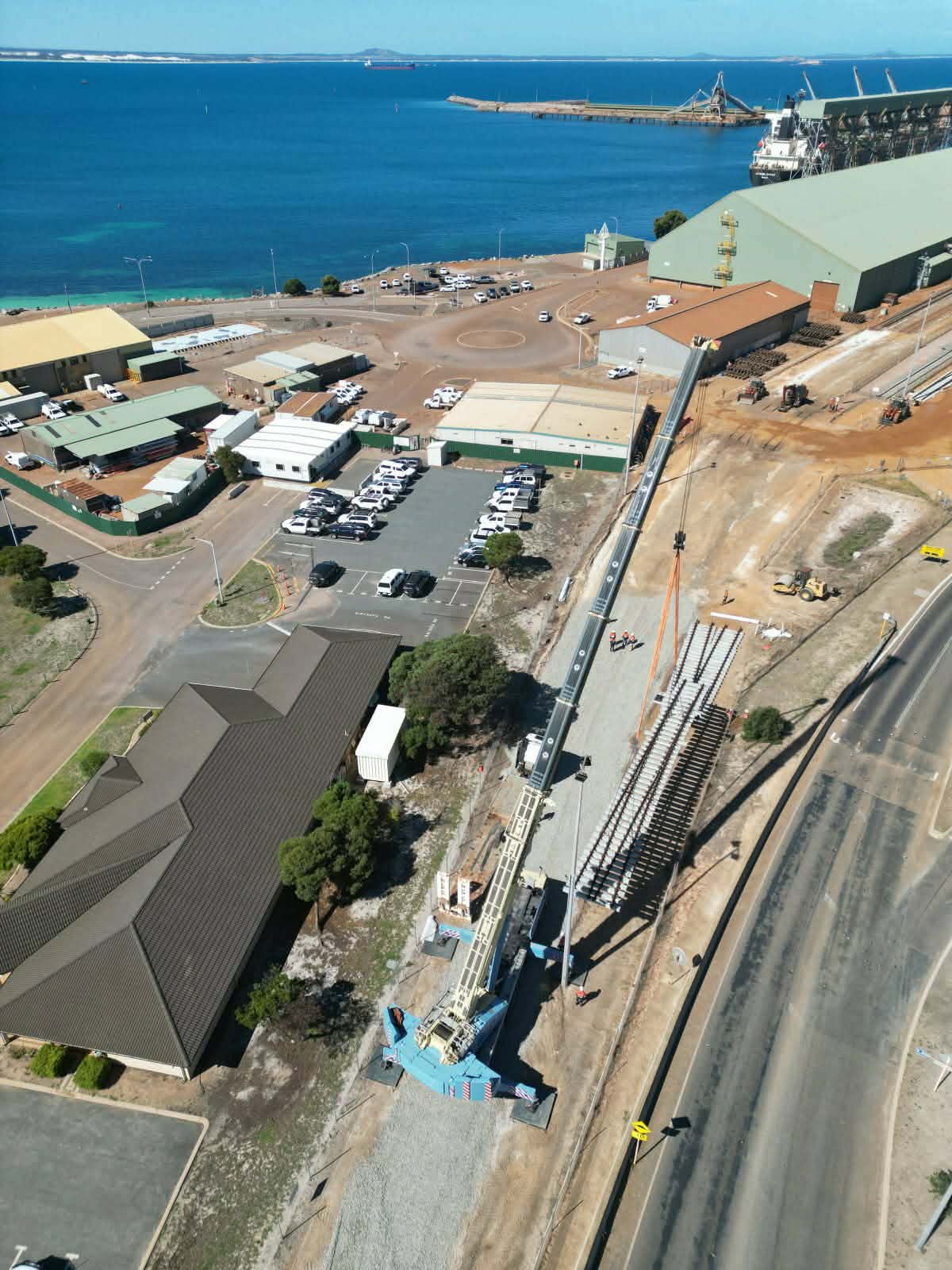Construction crane installing new rail tracks near a coastline with buildings, parking lot, and water in the background.