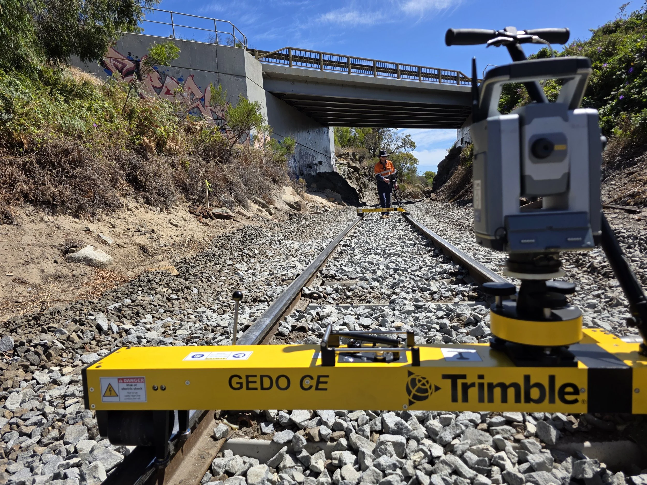 Railway track survey with surveying equipment and a worker measuring on the track, under a bridge, with trees and graffiti on the wall, sunny day.