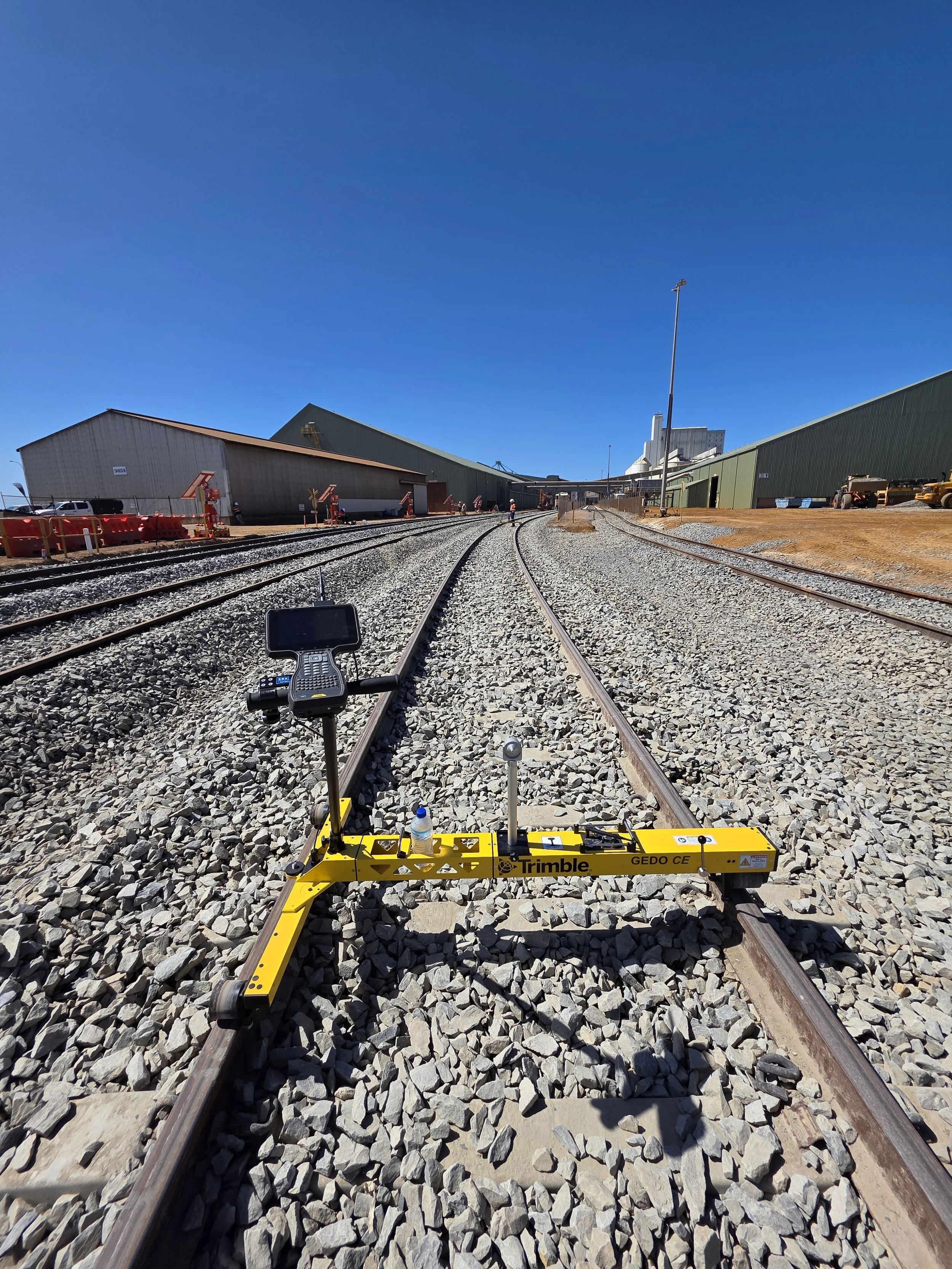 Railway tracks with a yellow surveying instrument labeled 'Trimble' on the gravel between the rails at an industrial railway yard.