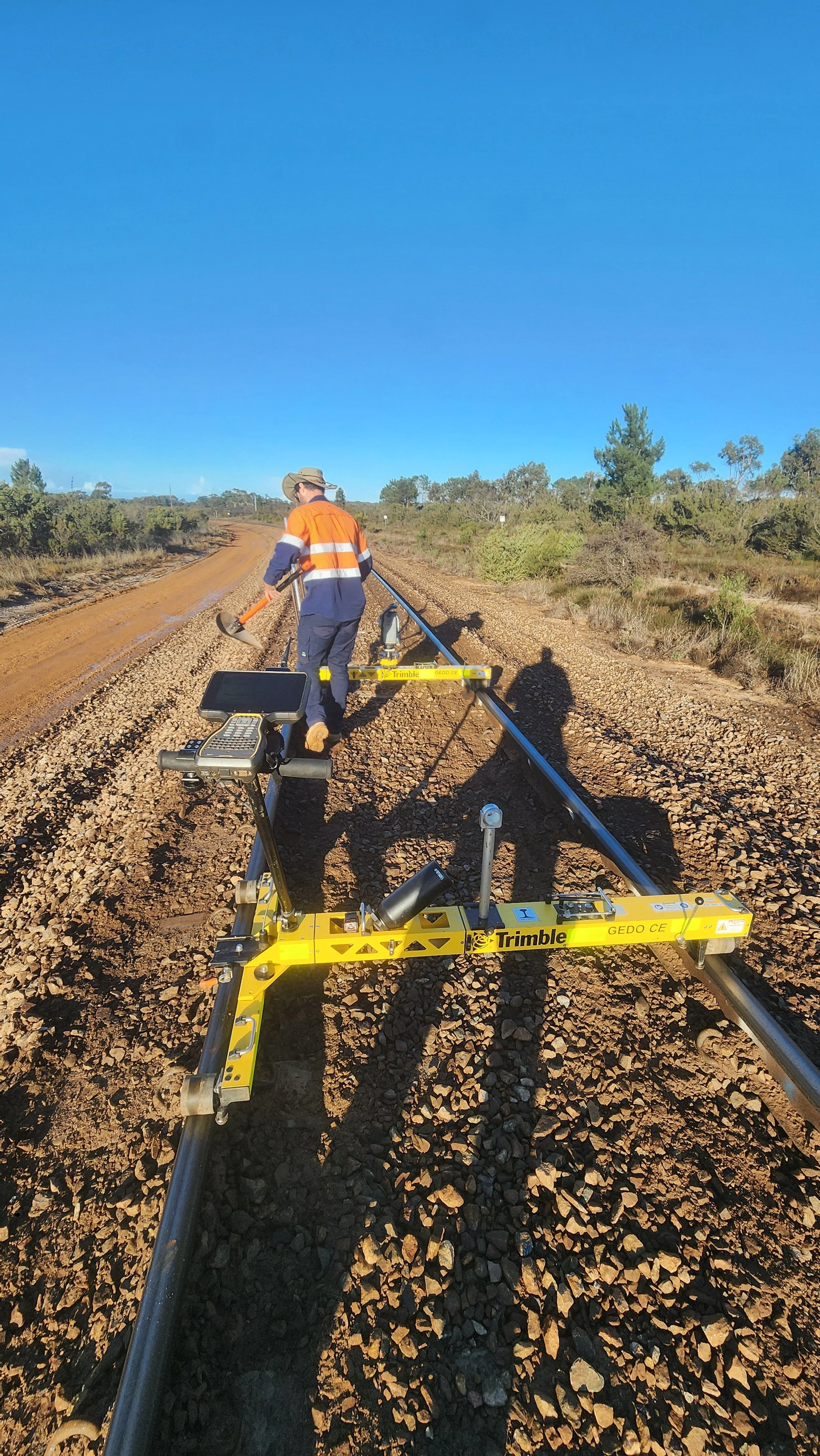 A surveyor on a dirt road using a trimble device for land measurement, wearing an orange and navy safety vest and a wide-brimmed hat, with shadows cast on the ground under a clear blue sky.