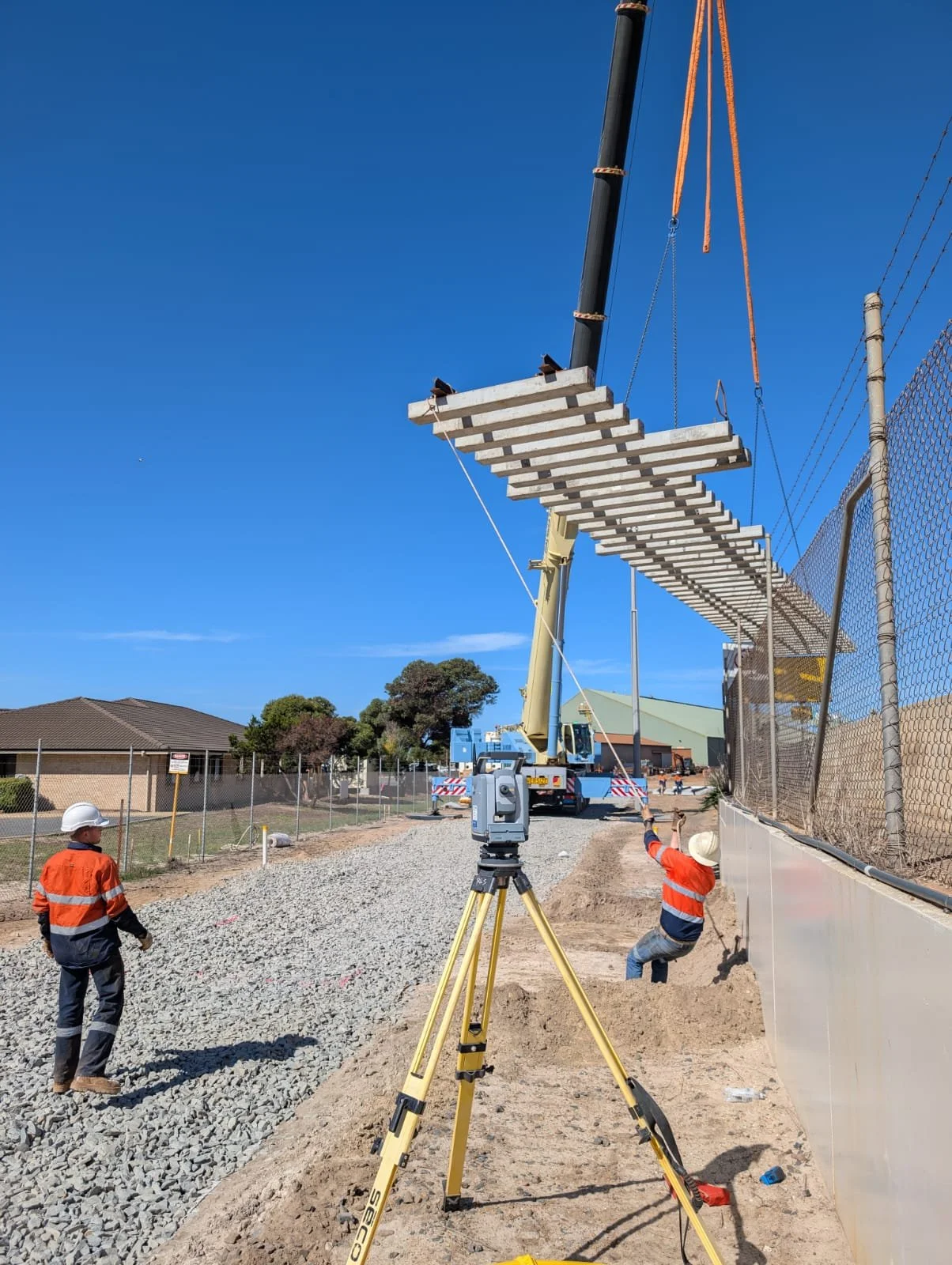 Construction workers on a construction site, with a crane lifting a wooden platform suspended in midair, and surveying equipment set up nearby on a sunny day.