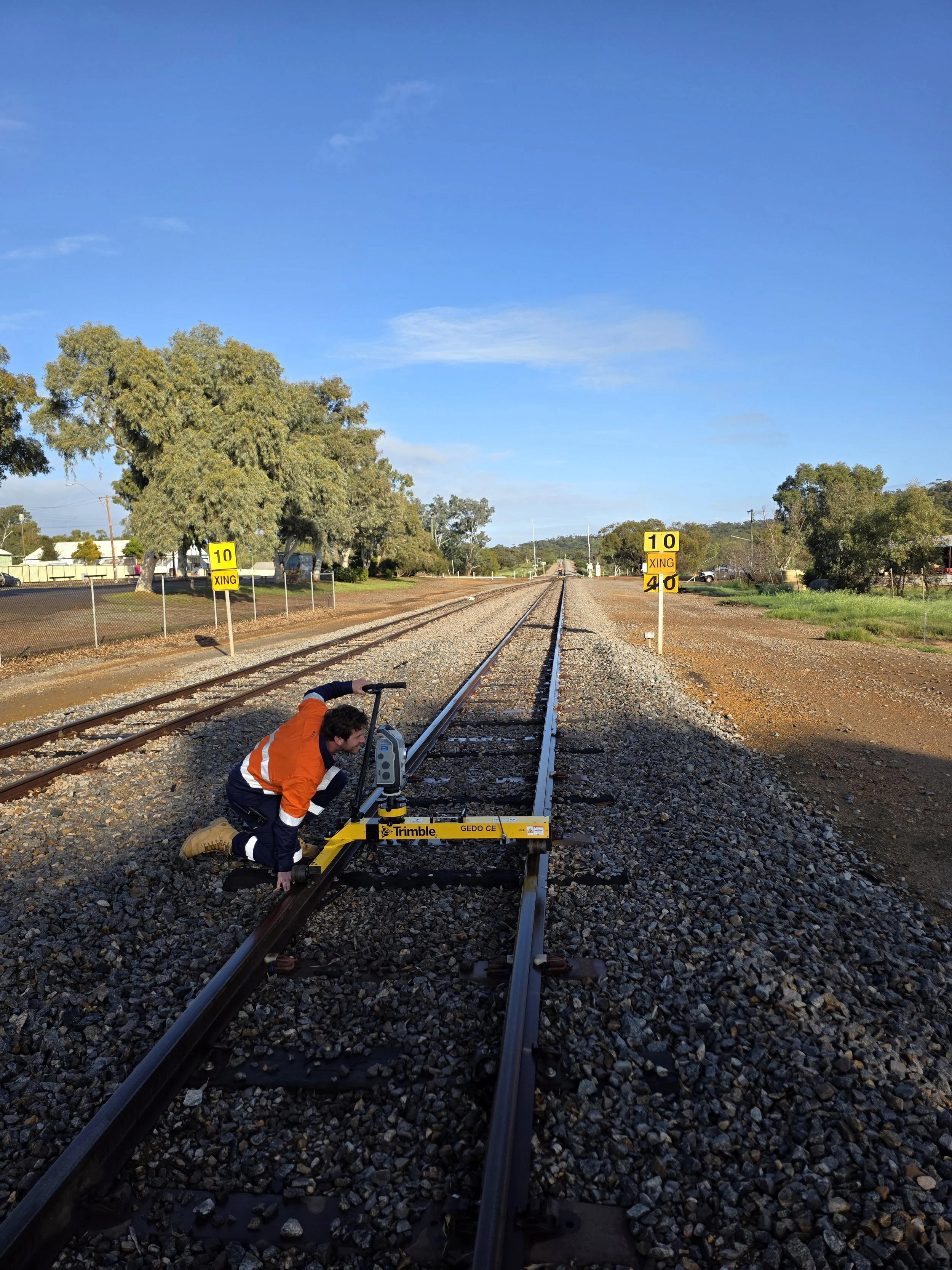 A worker in an orange safety jacket kneeling next to railroad tracks, inspecting or adjusting a piece of measuring equipment on the tracks. The scene is outdoors with trees and a clear blue sky, and yellow signs on either side of the tracks indicating speed limits and crossing information.