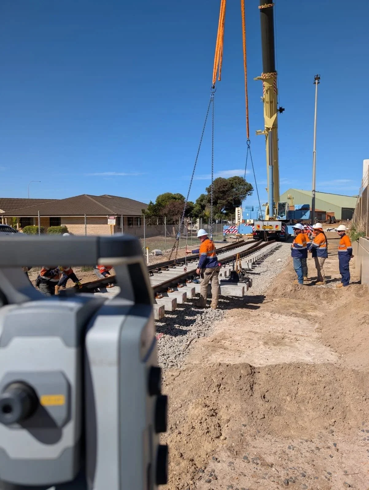 Construction workers in orange safety vests and white helmets working on a railway track with a crane lifting railroad ties on a sunny day.