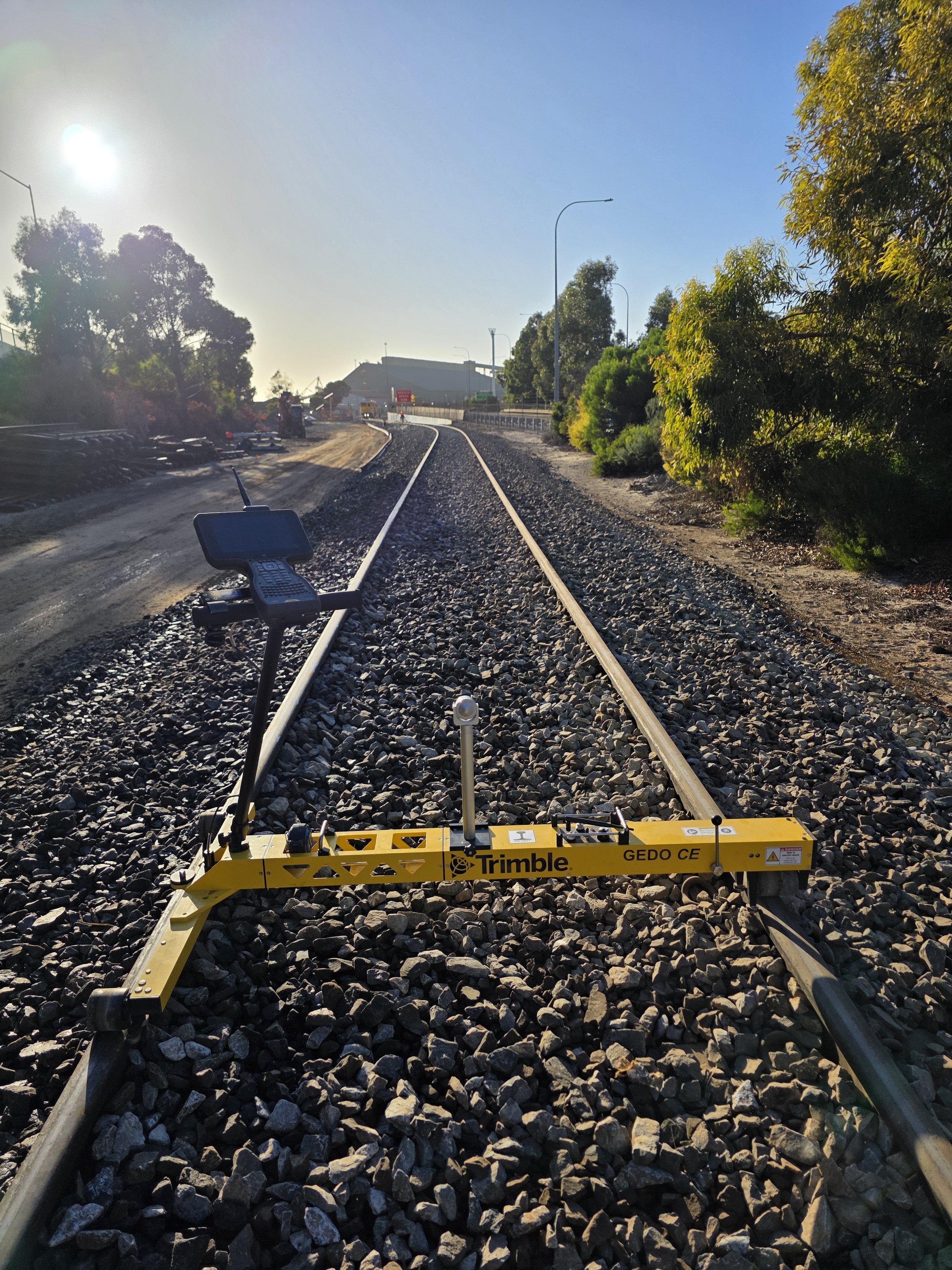 Railroad tracks under construction with survey equipment and construction site in the background, trees on the right side, and a bright sun in the sky.