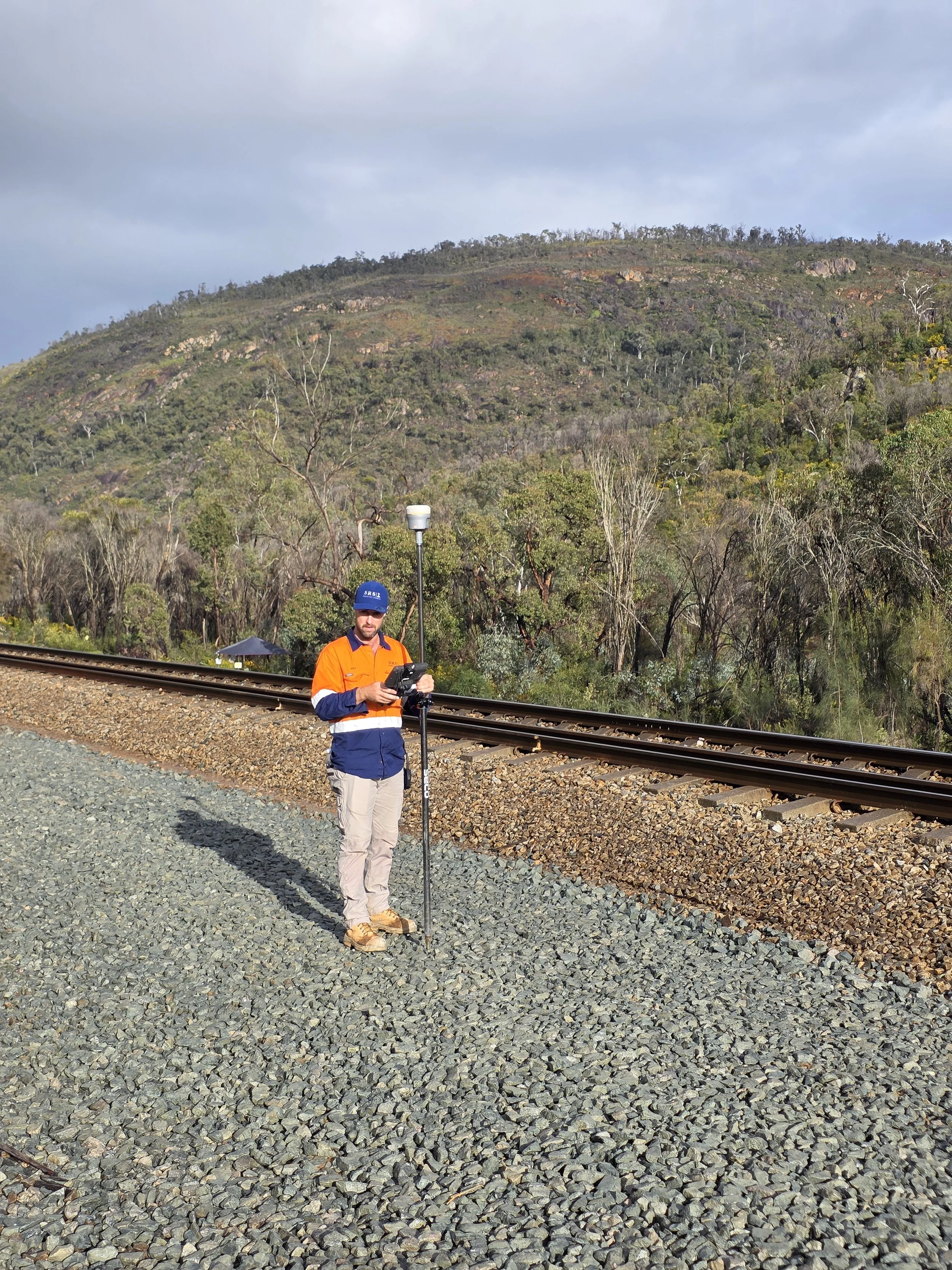 A man in an orange and blue jacket is standing beside train tracks on a gravel surface, holding a surveying instrument, with a mountainous landscape and trees in the background.