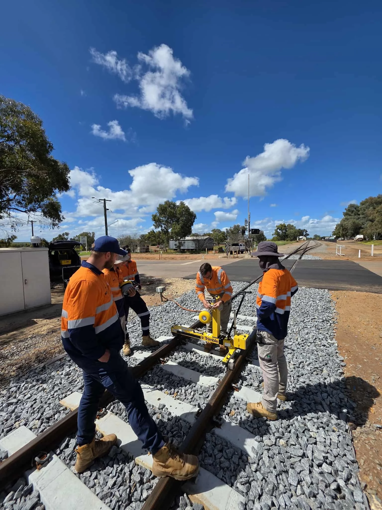 Workers in orange high-visibility jackets and boots installing and adjusting a railway track under a blue sky with scattered clouds.