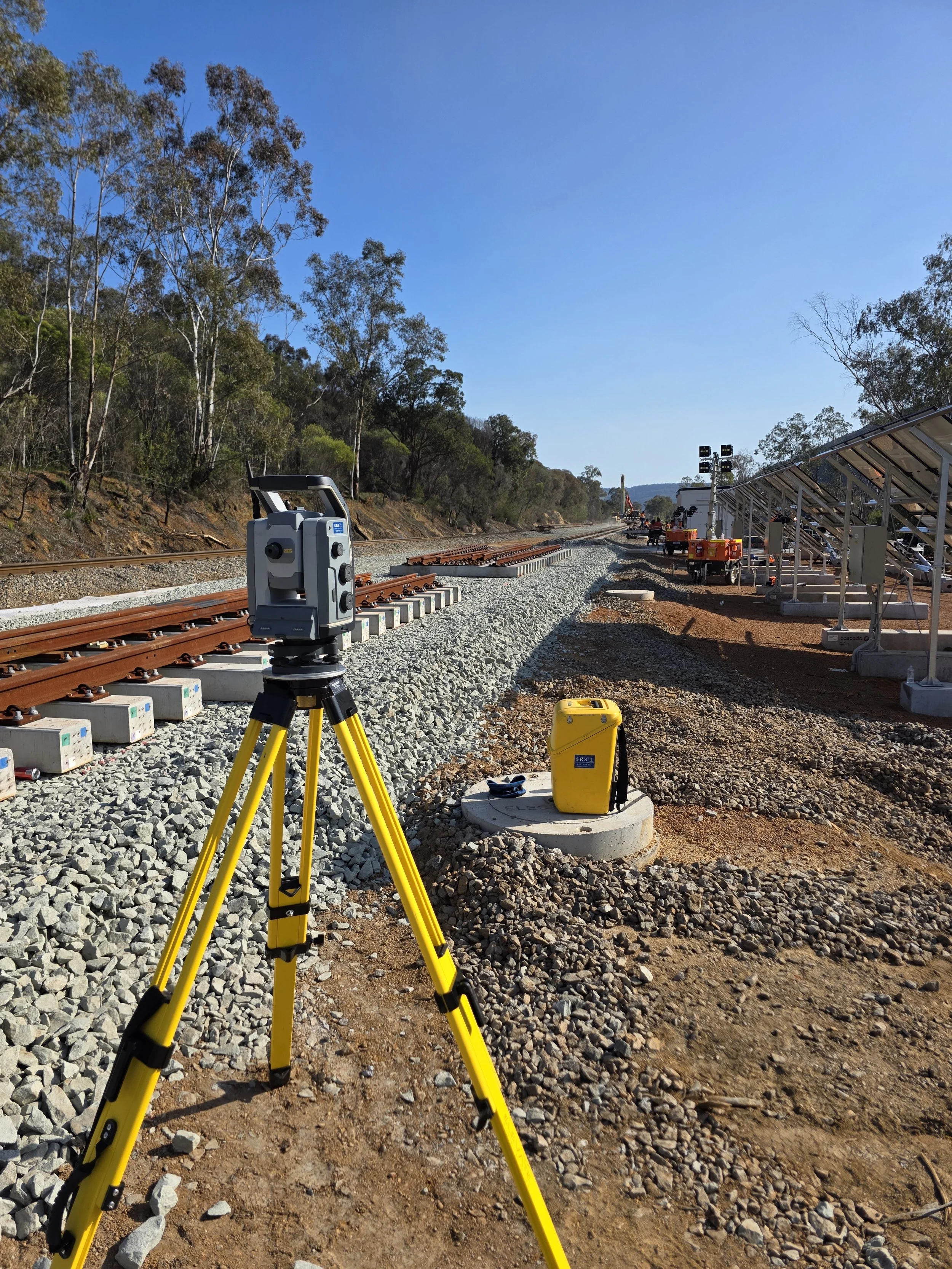 Construction site with railway track, surveying equipment on tripod, solar panels, and construction workers in the background under a clear blue sky.