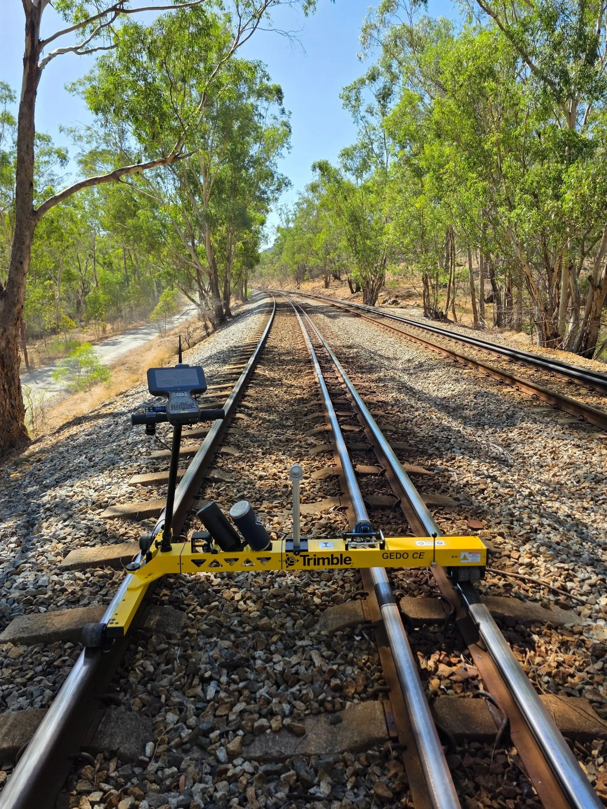 A railway track with a yellow surveying device labeled 'Trimble GEDO CE' positioned on the tracks. The scene is surrounded by trees and has a clear blue sky above.