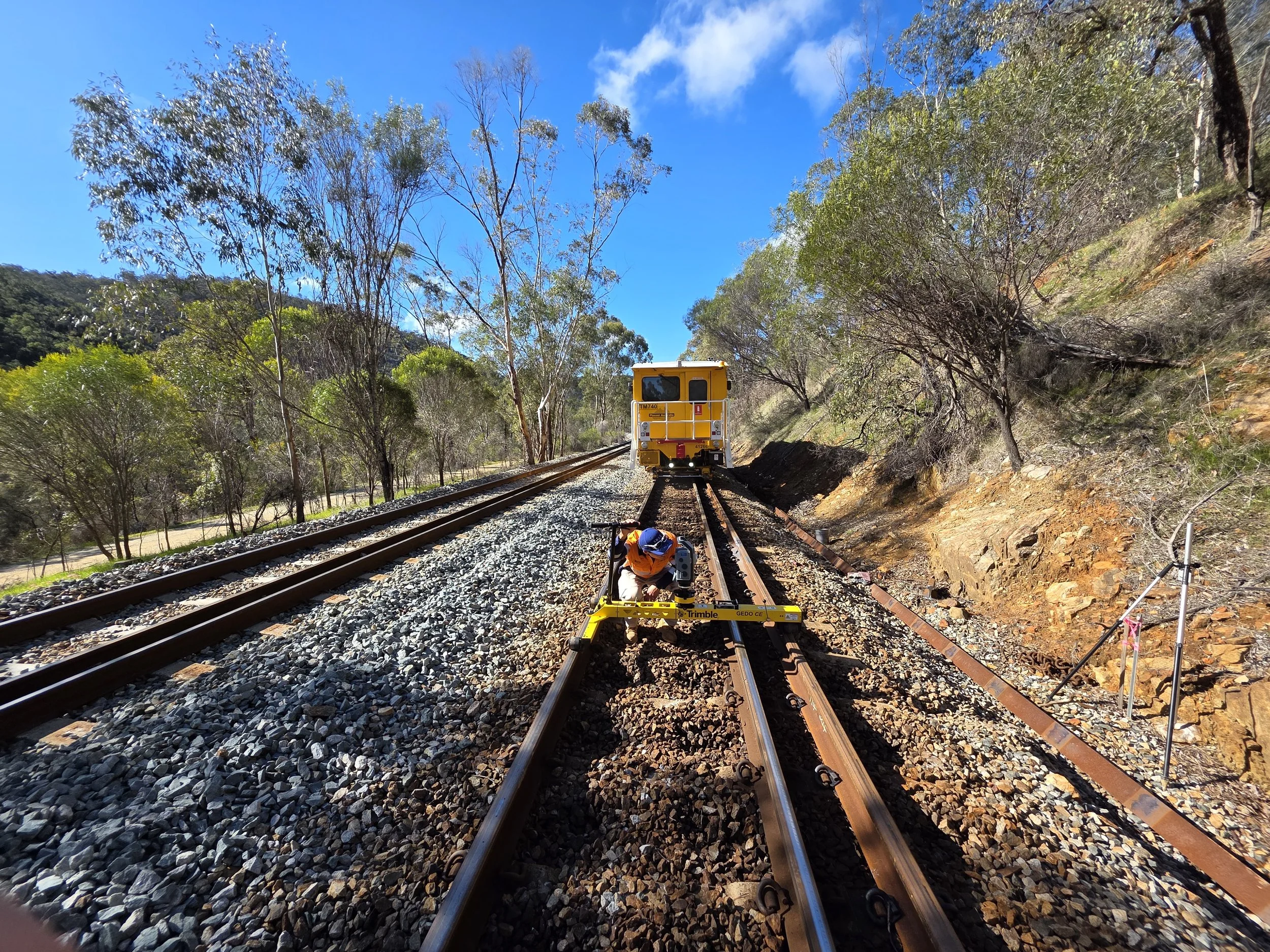 A worker in safety gear uses a level on railroad tracks, with a maintenance vehicle approaching on the same tracks, set in a wooded outdoor area on a sunny day.