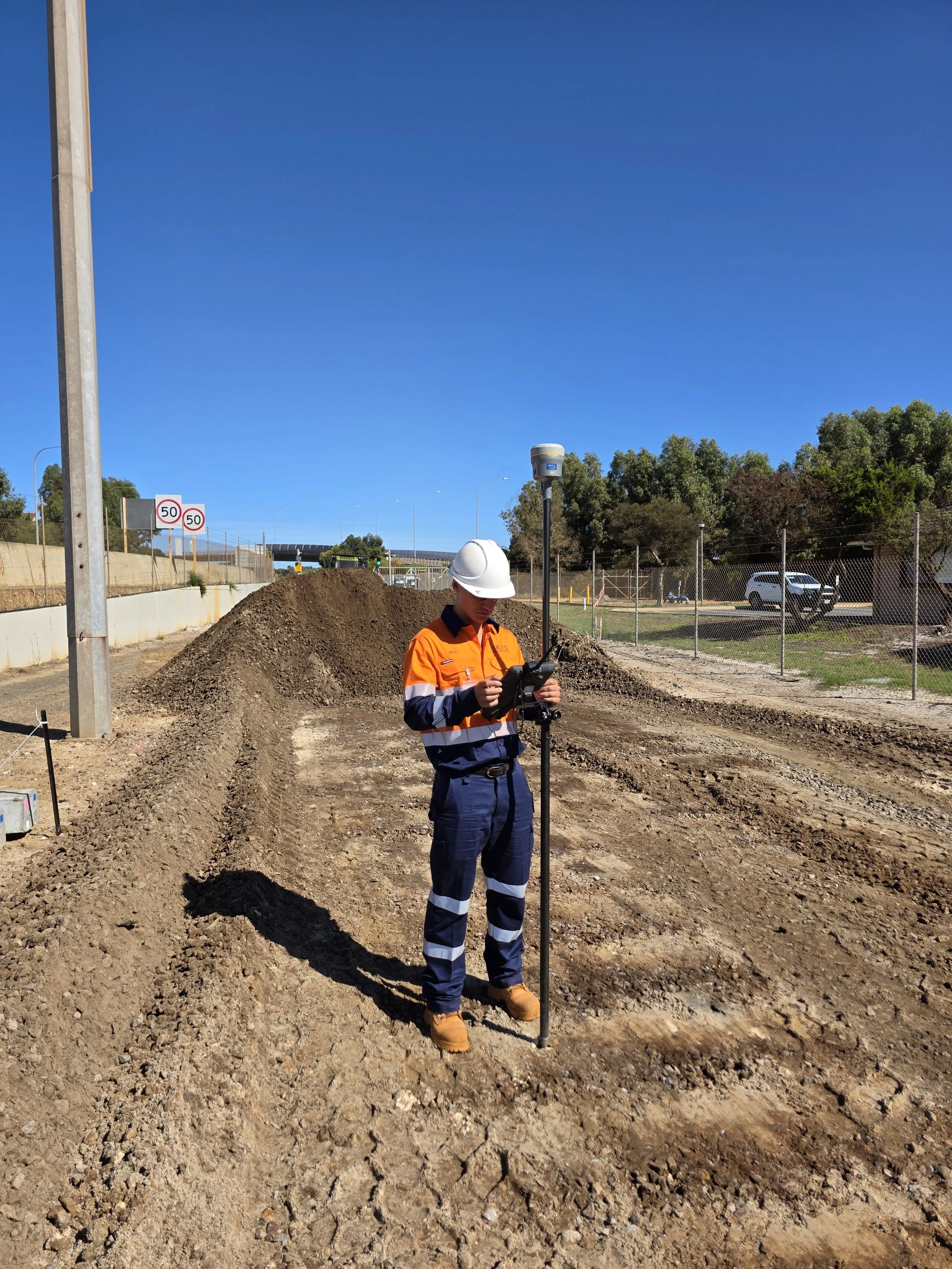 A construction worker wearing an orange safety vest and white hard hat using surveying equipment at a construction site on a clear, sunny day.