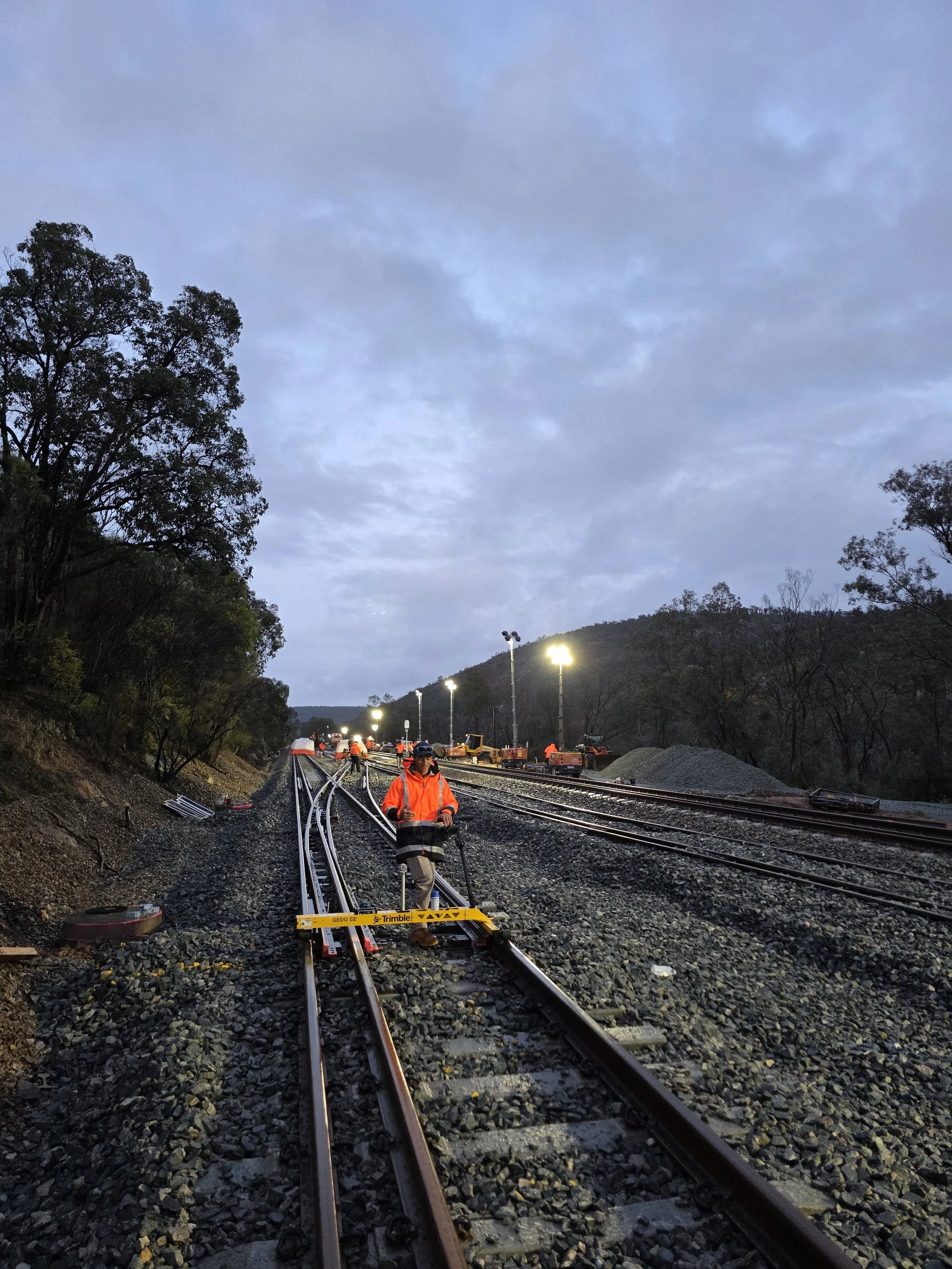 Railroad workers in high-visibility orange jackets working on train tracks at dusk, with construction equipment and bright lights in the background across a hilly landscape.