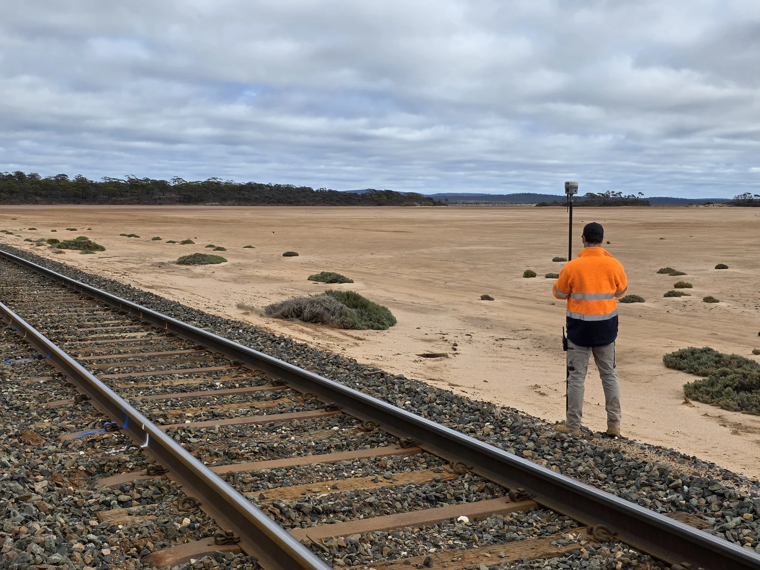 A person wearing an orange safety jacket and gray pants stands next to a railway track, looking across a desert-like landscape with sparse bushes and a cloudy sky.