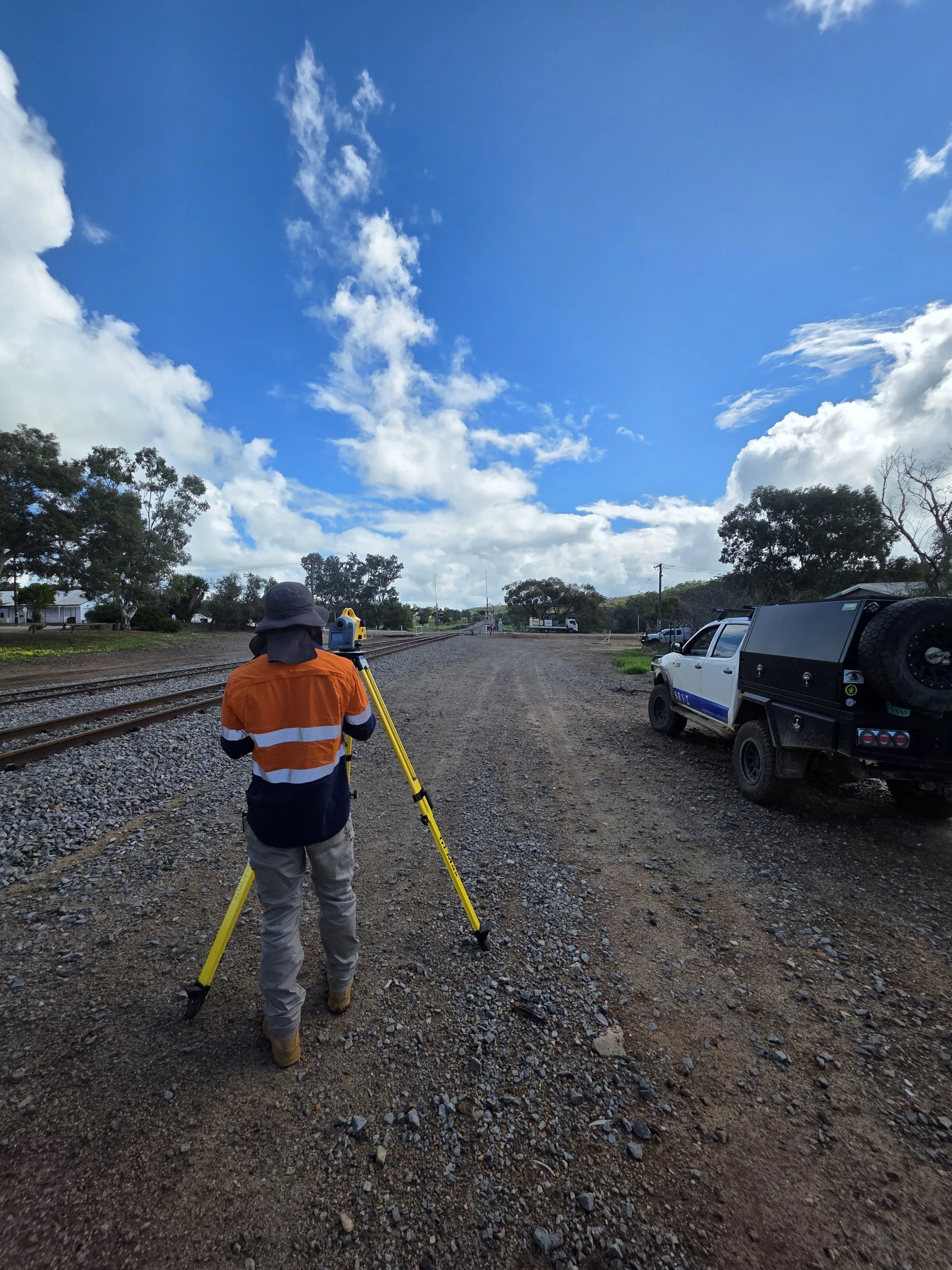 A surveyor in an orange and navy safety vest using a leveling instrument on a tripod near railway tracks on a gravel path. A pickup truck is parked nearby under a blue sky with scattered clouds.