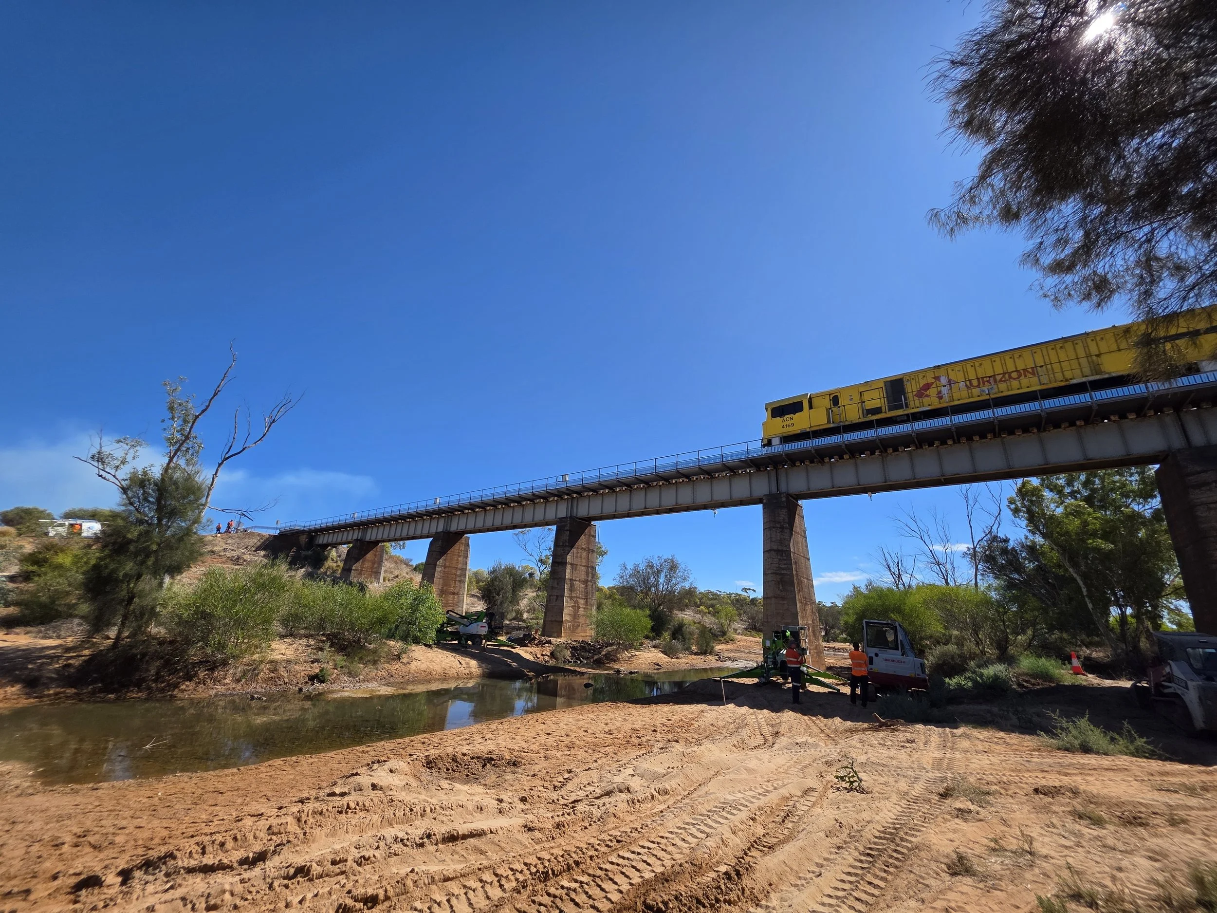 A yellow train crossing a bridge over a small river in a desert area with construction workers working on the riverbank.