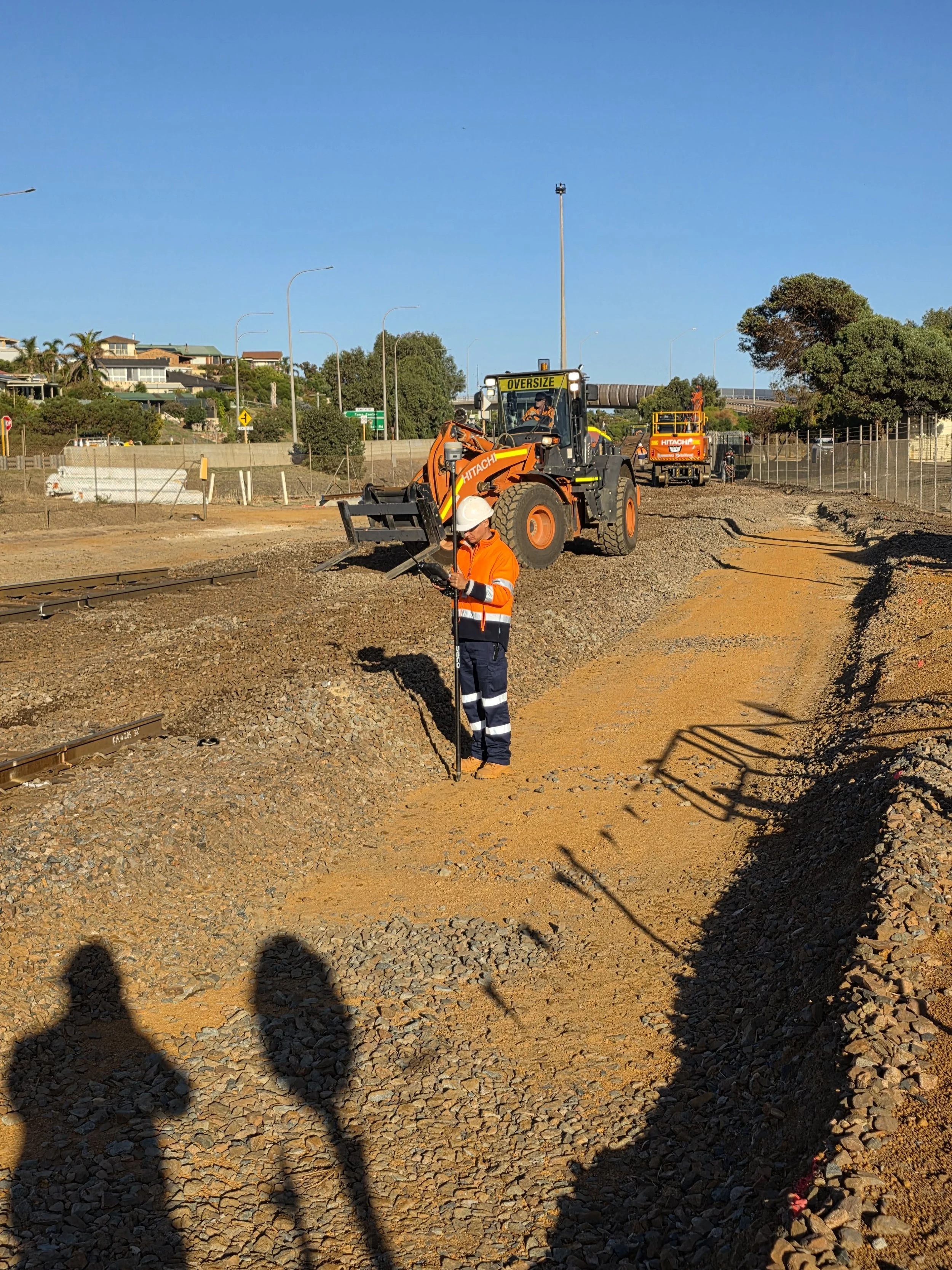 Construction site with two workers and heavy machinery, including a large orange loader, working on a dirt and gravel pathway next to railroad tracks under a clear blue sky.