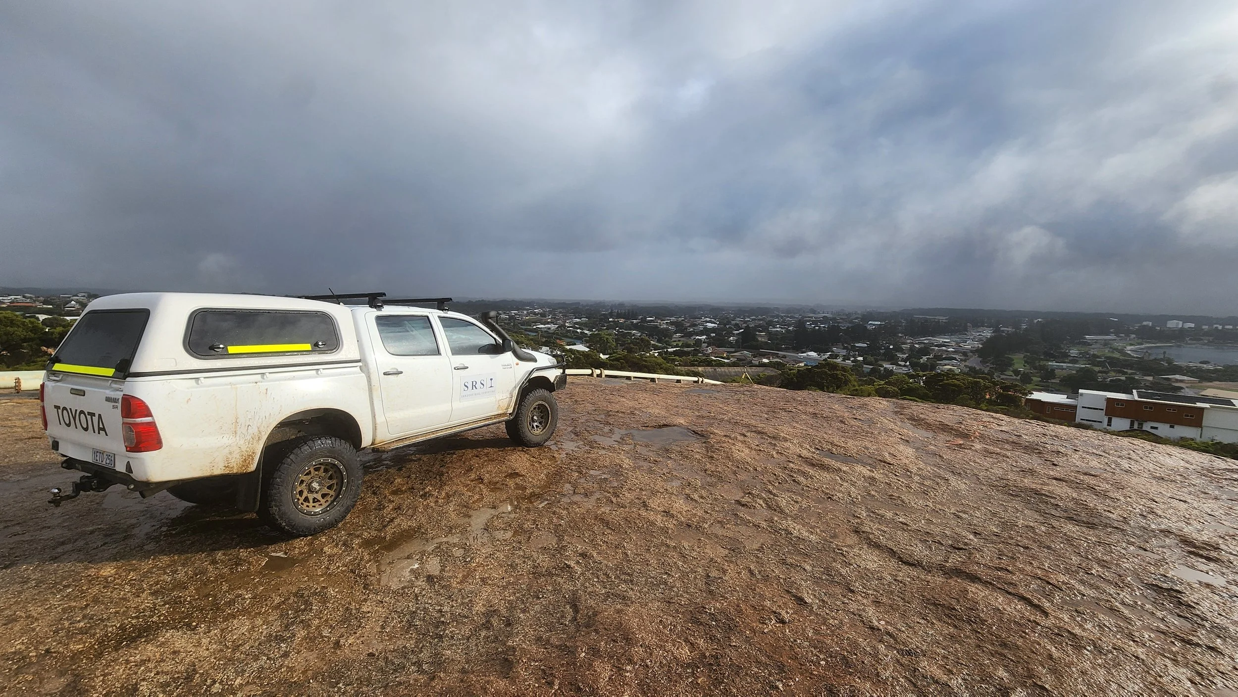 White Toyota pickup truck with a canopy parked on a rocky hilltop overlooking a cityscape under cloudy sky.