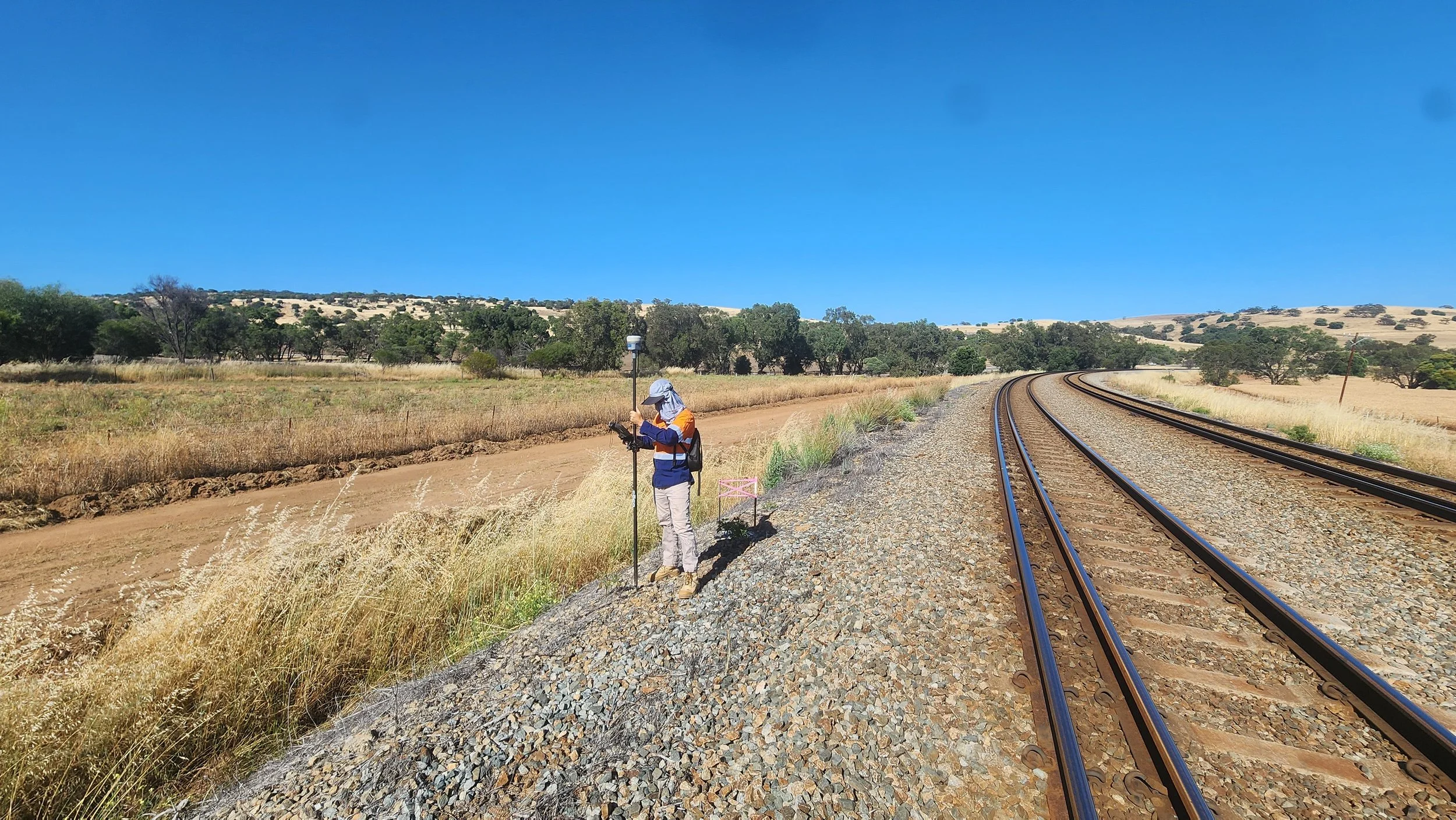 A person wearing outdoor clothing and a sun hat standing beside a railway track in a rural landscape, using a surveying instrument.