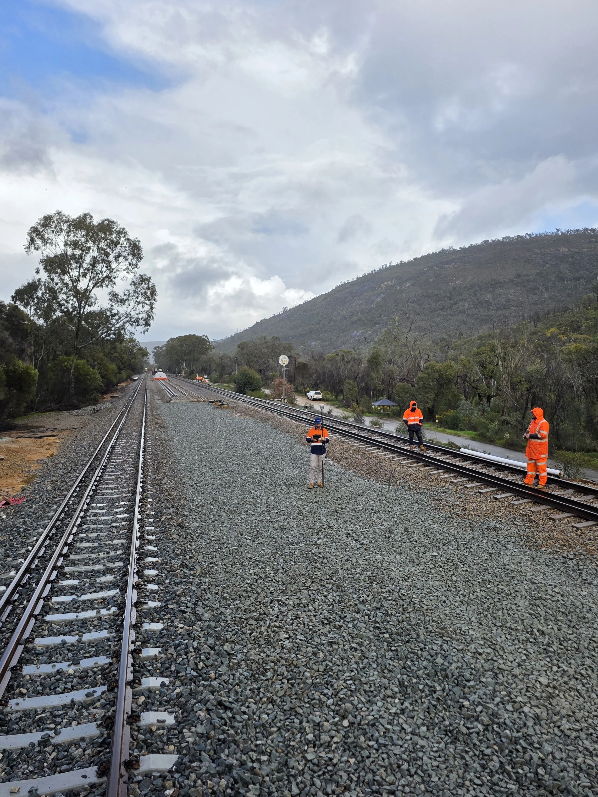 Three workers in orange safety gear inspecting railway tracks in a rural area with trees and hills in the background under cloudy sky.