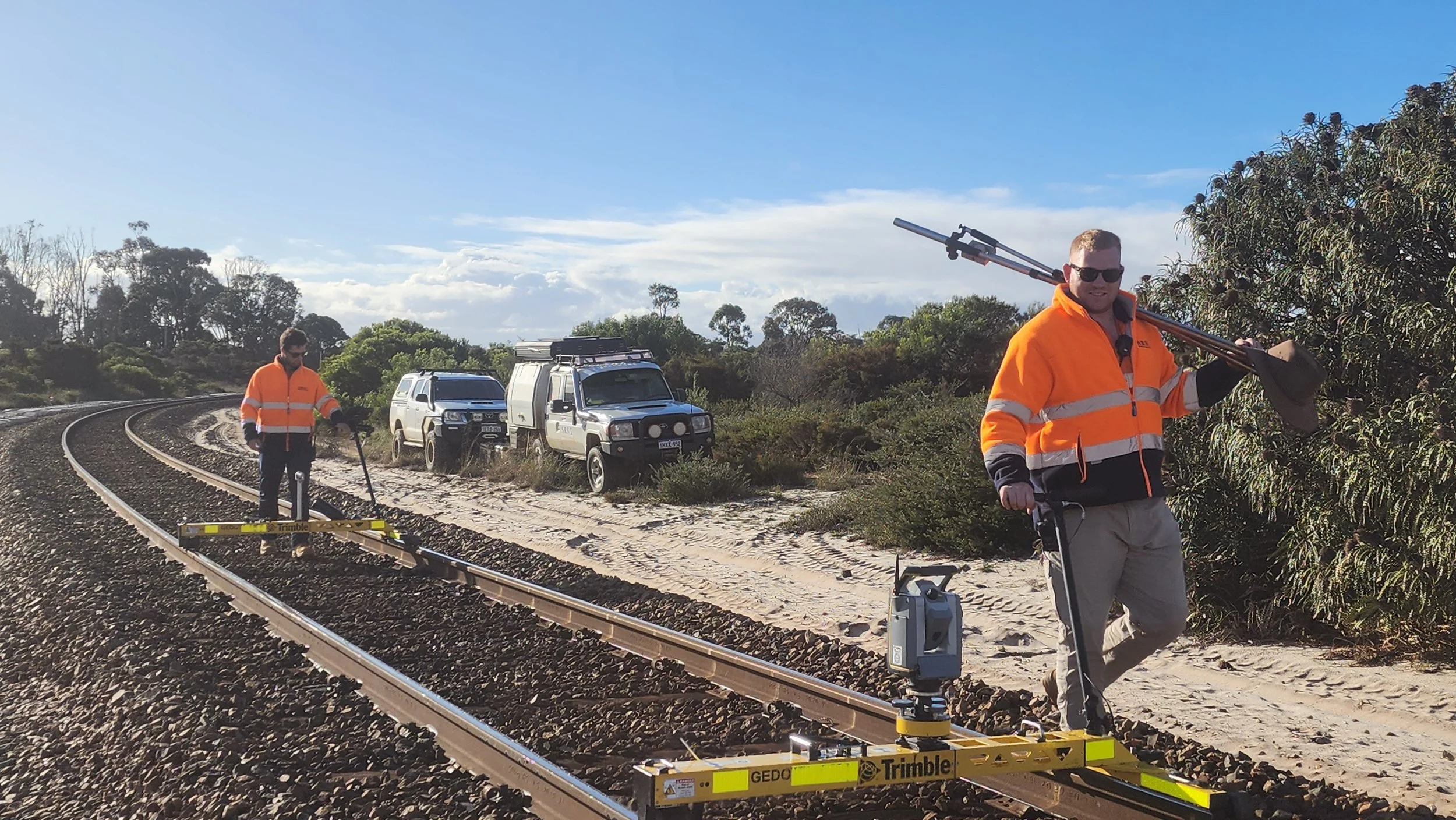 Two surveyors in orange jackets working on railroad tracks near a forest, with vehicles parked on a dirt path beside the tracks.