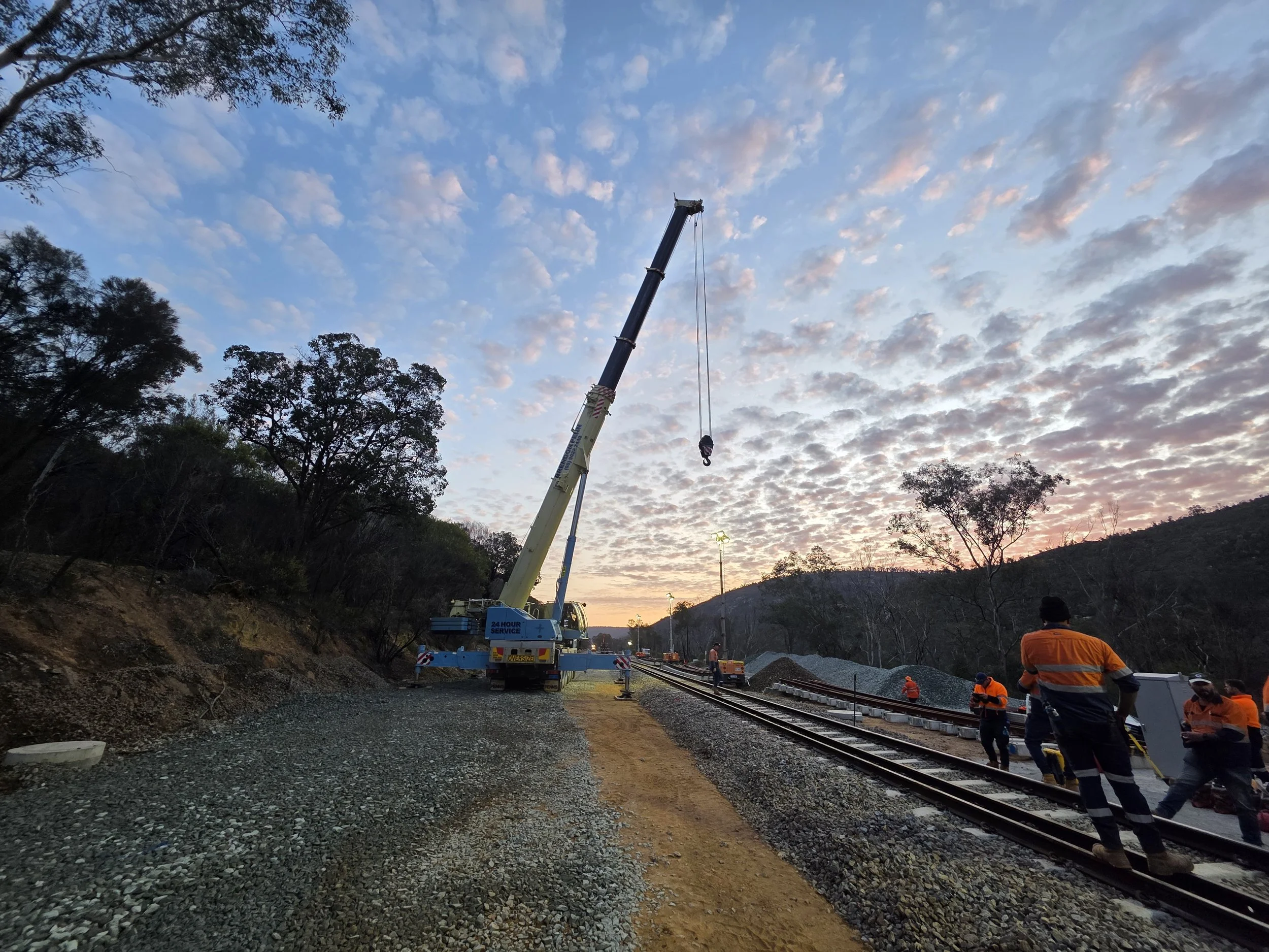 Construction workers working on railway tracks at sunset with a crane lifting a hook, and trees in the background.