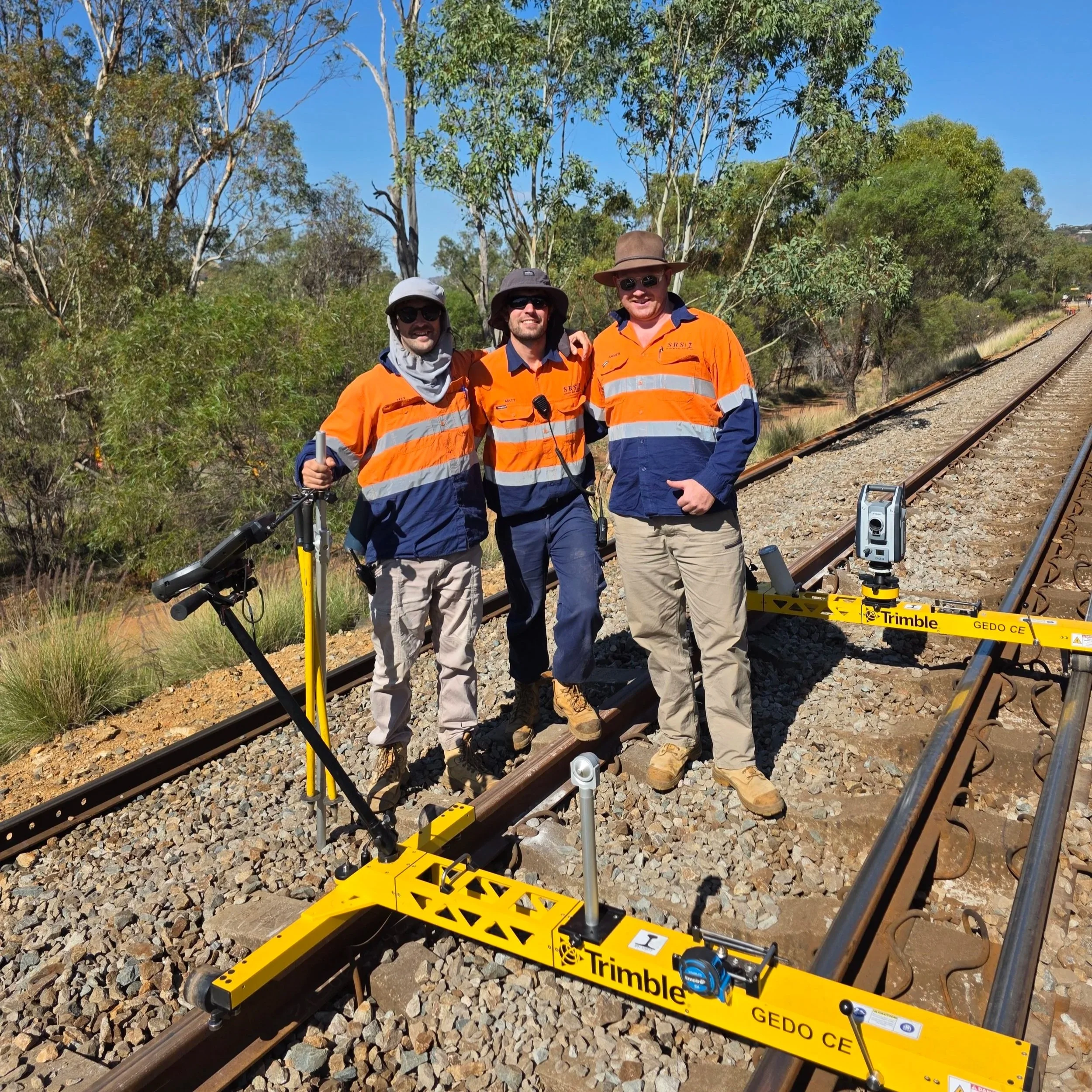 Three men in orange and navy high-visibility jackets standing on railway tracks in a rural area with trees and blue sky, using surveying equipment.