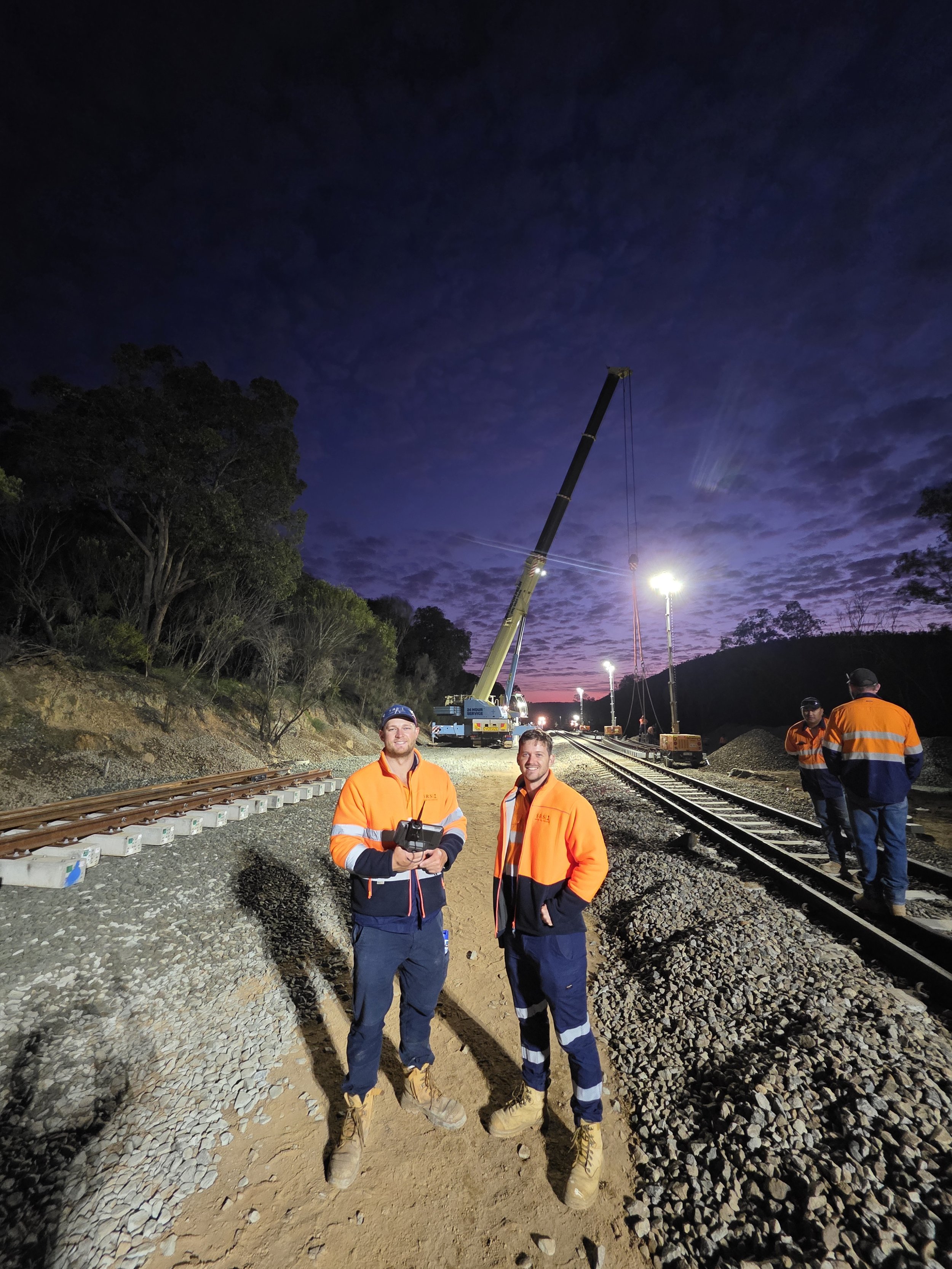 Four workers in orange safety gear standing on train tracks at dusk, with a crane lifting equipment in the background and a colorful sky.