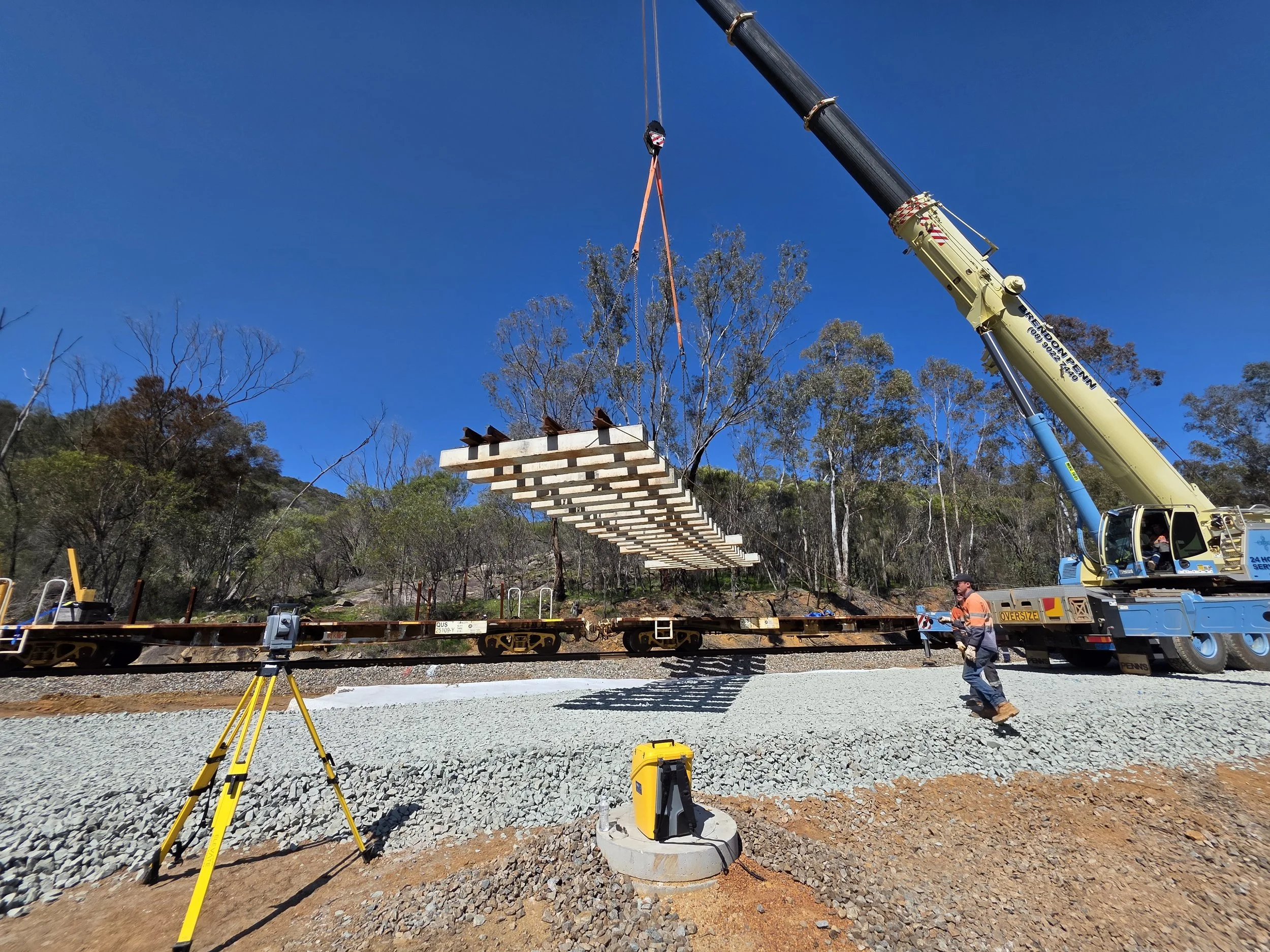 A crane lifting wooden railroad ties onto train tracks at a construction site with trees and a clear blue sky in the background.