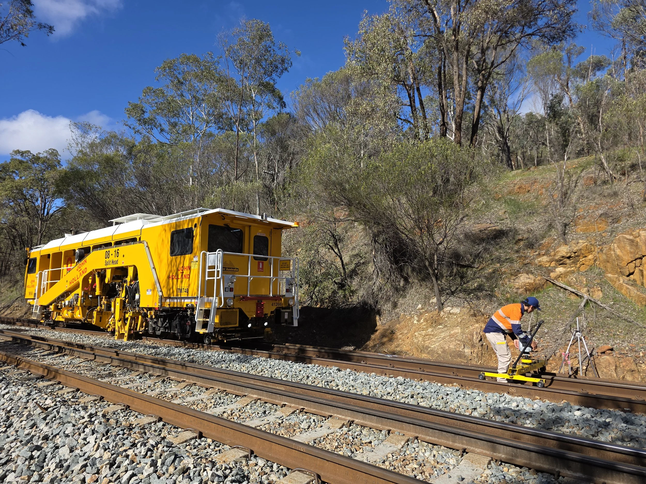 A worker in orange and navy safety gear operating a precision measurement device on railway tracks near a yellow maintenance vehicle in a rural outdoor setting with trees and rocks.