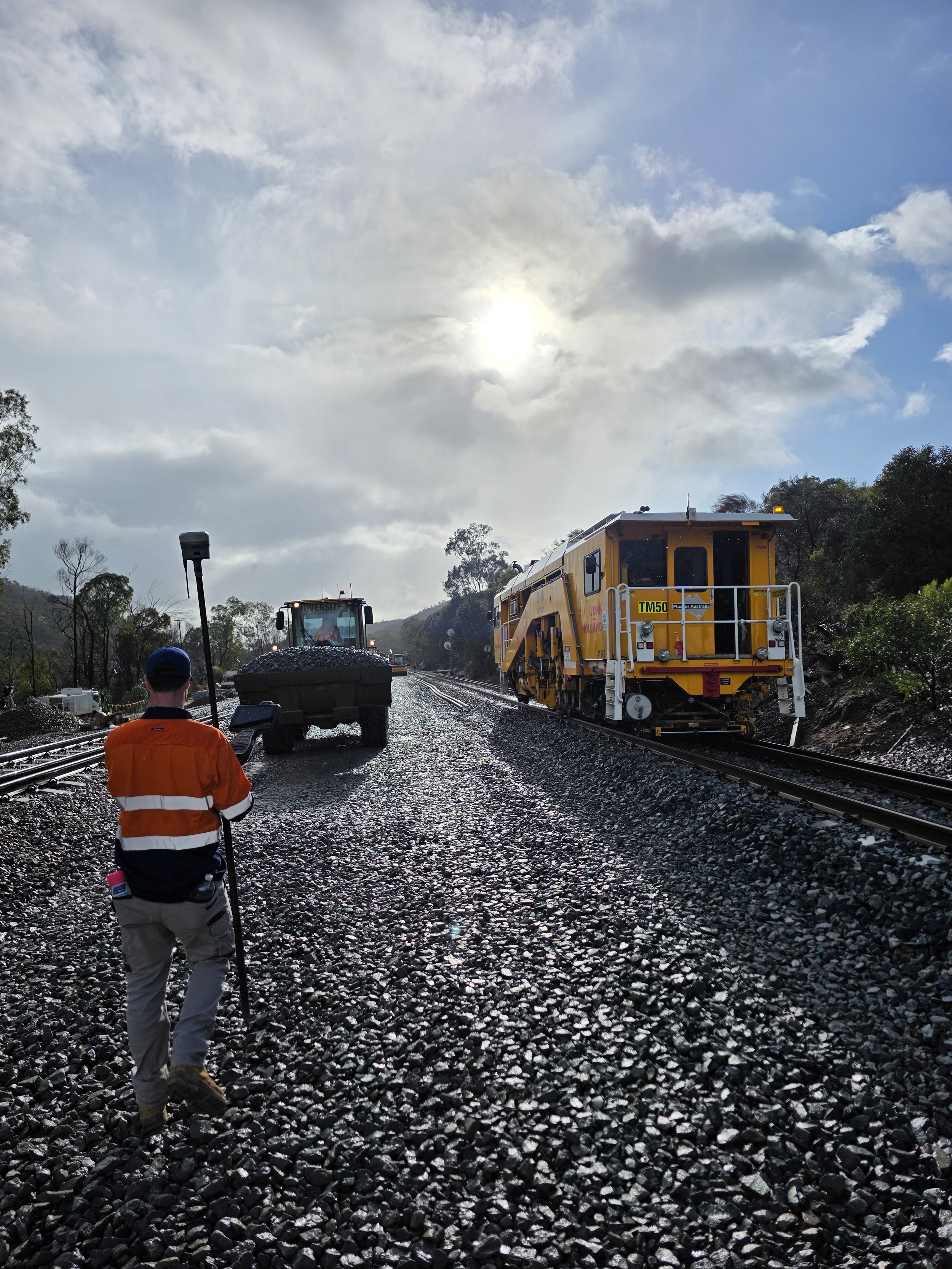 A railway worker in an orange safety vest and gray pants walking on a gravel train track near a yellow rail maintenance machine, with another vehicle and a pole with equipment in a rural area under a cloudy sky.