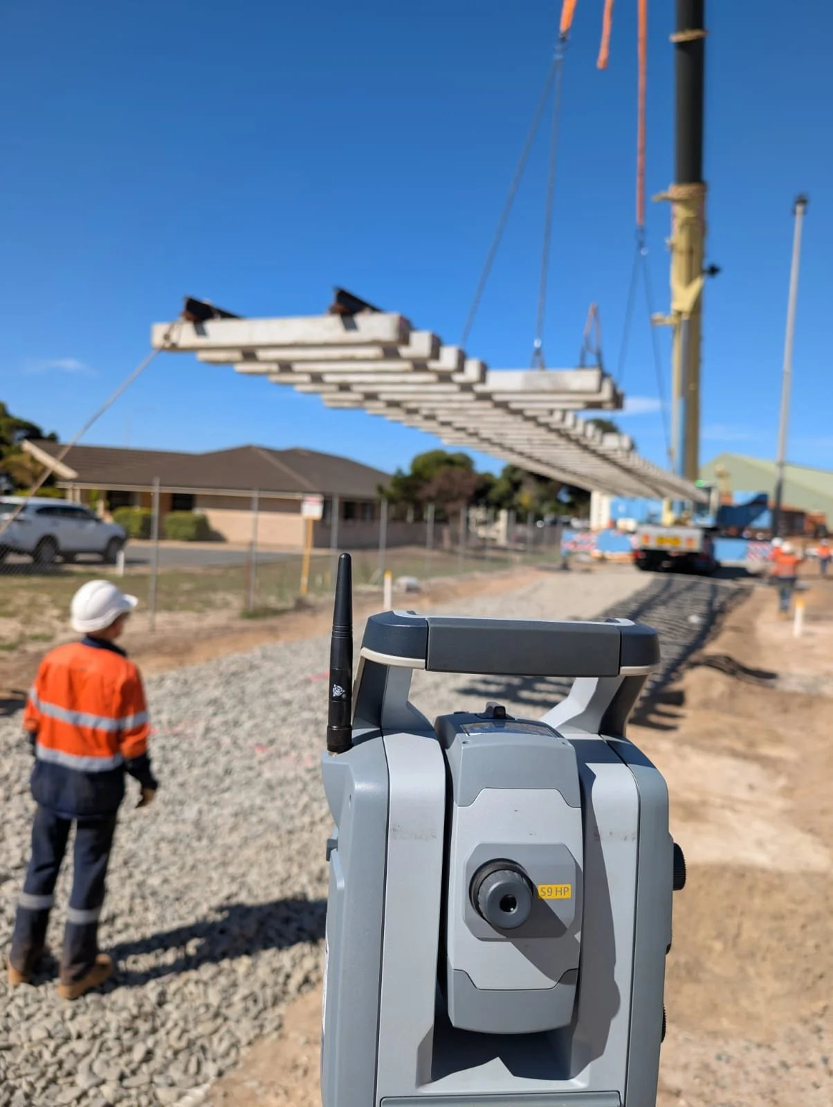 A construction worker in an orange safety vest and white hat standing on a gravel path. In the foreground, a surveying instrument is visible, with a yellow label marked 'S9 HP'. In the background, a crane is lifting long concrete beams, with a clear blue sky overhead.
